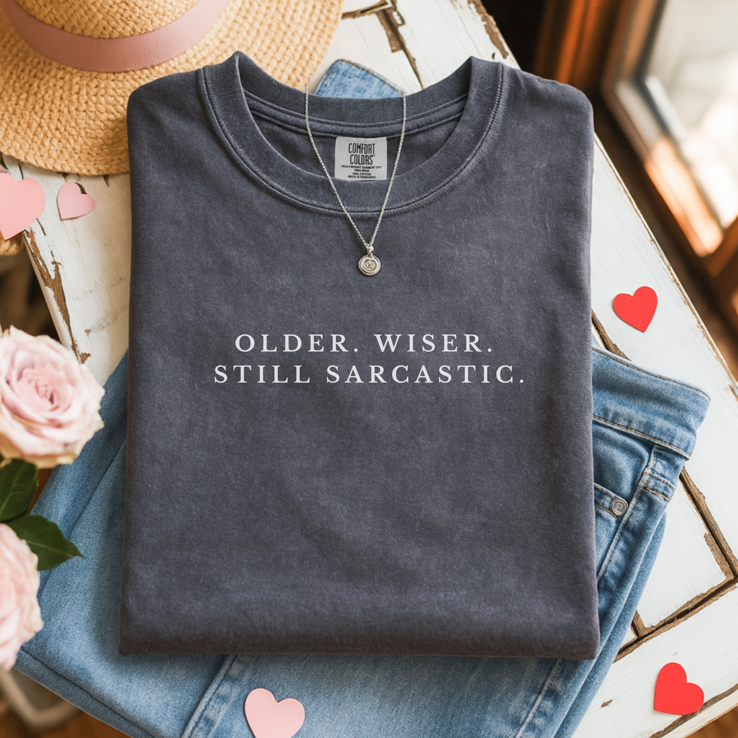 Folded dark gray shirt with 'OLDER. WISER. STILL SARCASTIC.' text, surrounded by flowers and a hat.