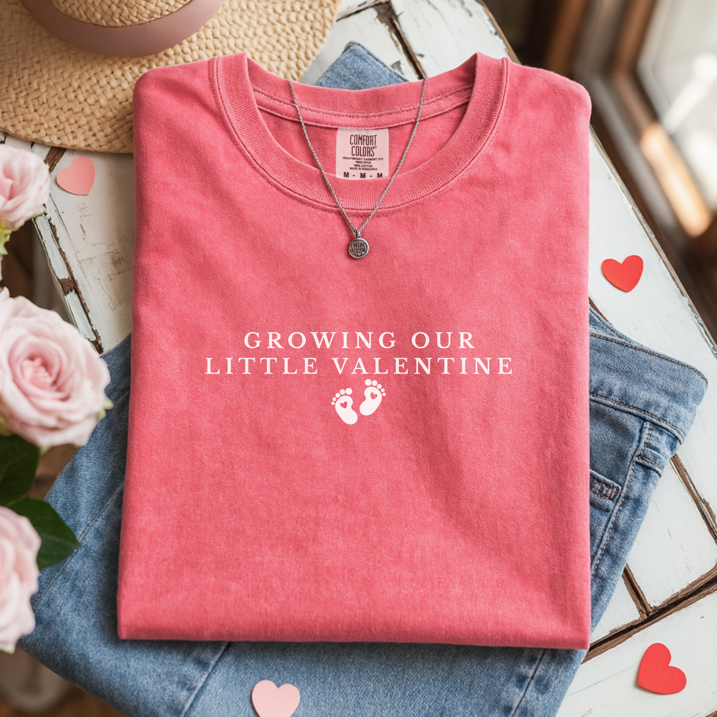 Folded pink t-shirt with 'Growing Our Little Valentine' text on a denim surface with flowers and a hat.