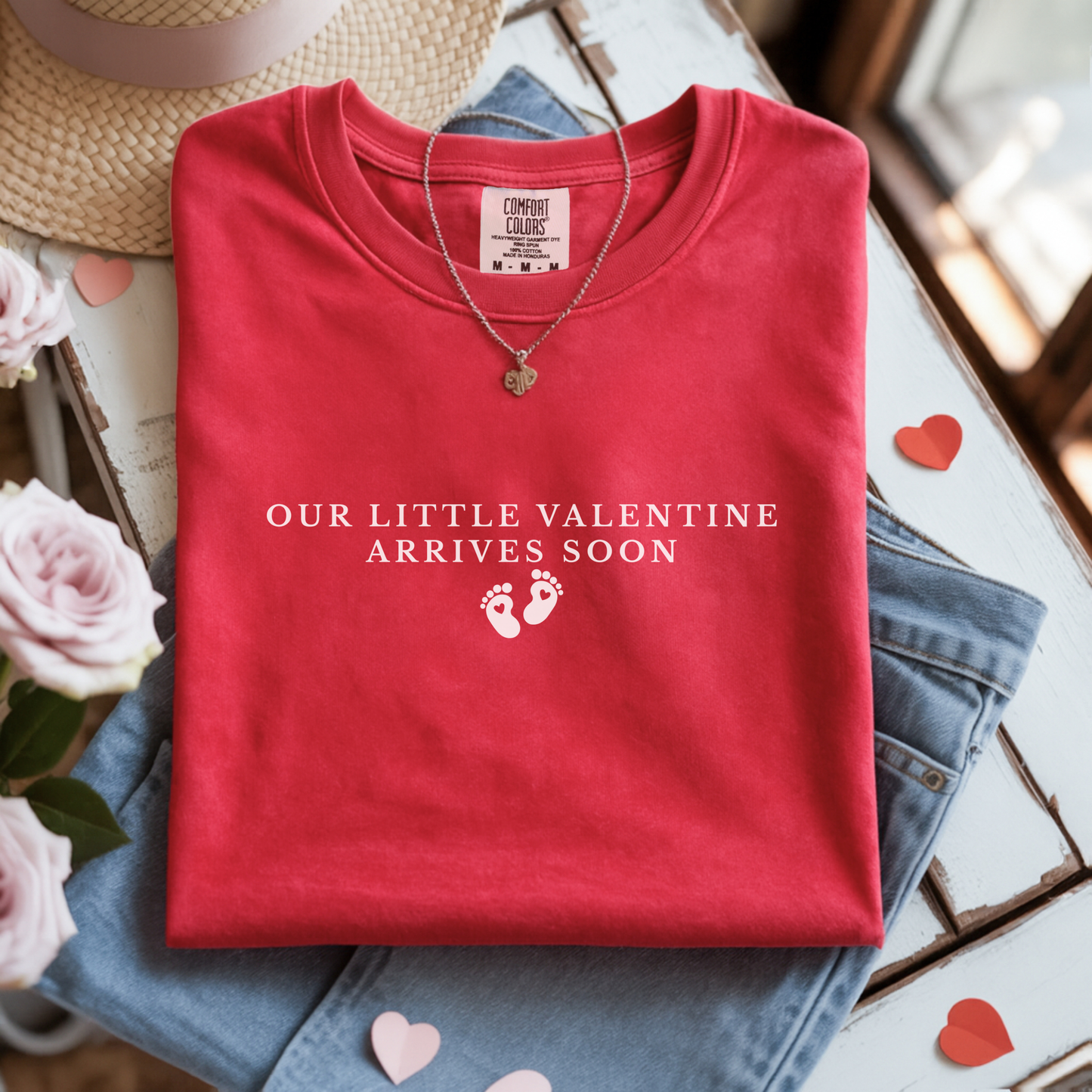 Folded red shirt with 'Our Little Valentine Arrives Soon' text on a surface with flowers and jeans.