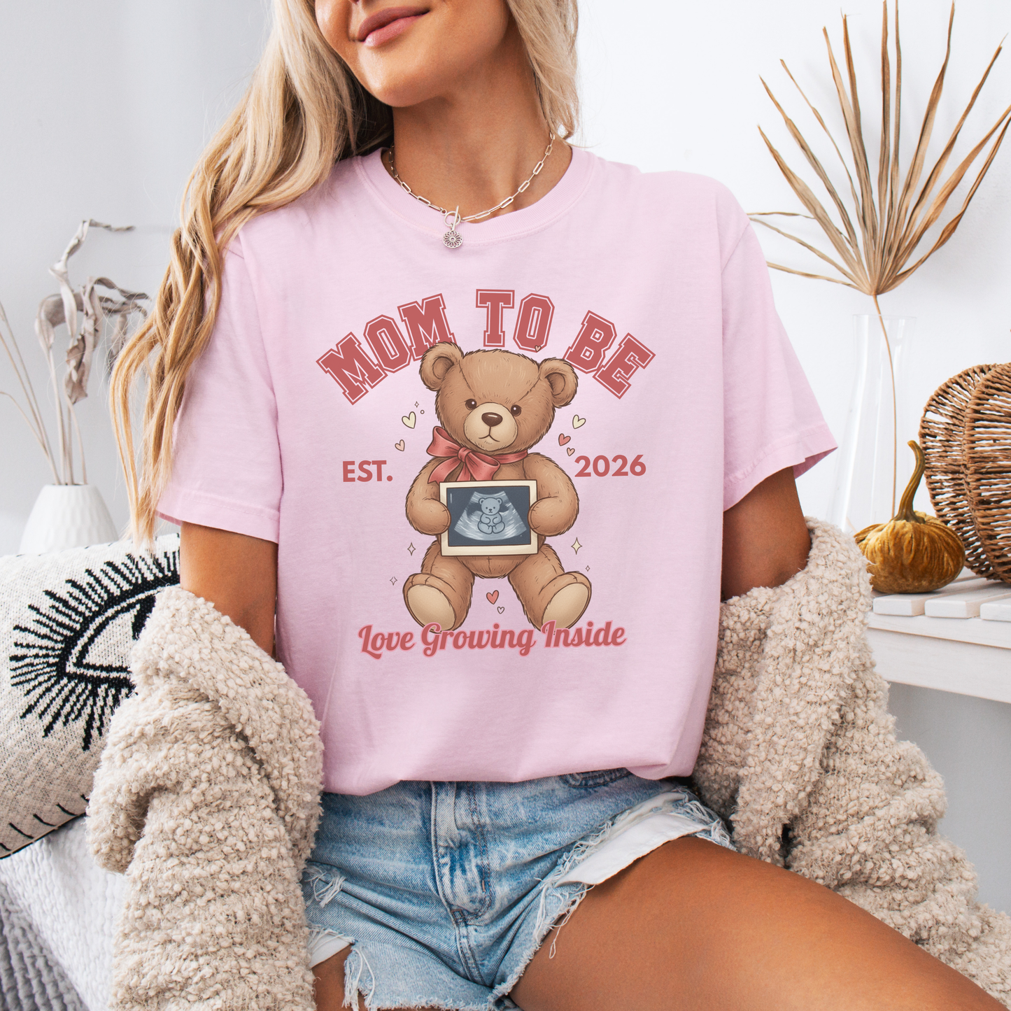 Woman wearing a pink 'Mom to Be' t-shirt with a teddy bear graphic, sitting in a cozy room.