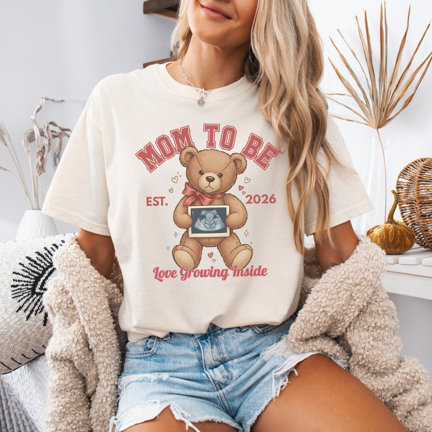 Woman wearing a 'Mom to Be' t-shirt with a teddy bear graphic, sitting on a couch.