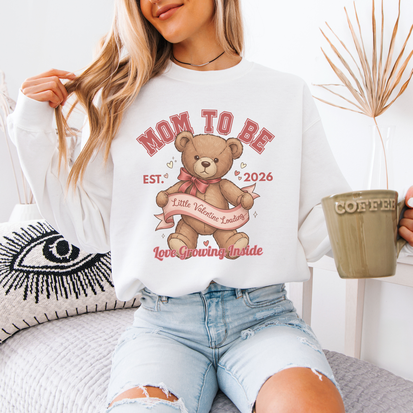 Woman wearing a 'Mom to Be' sweatshirt with a teddy bear graphic, holding a coffee mug.