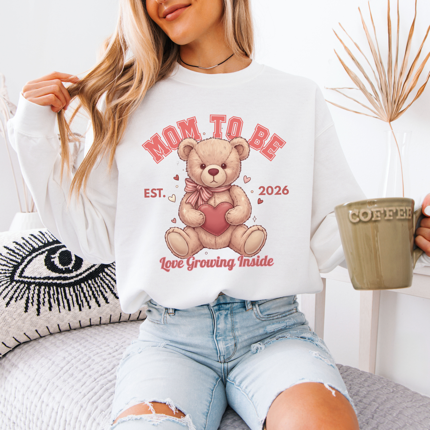 Woman wearing a 'Mom to Be' sweatshirt with a teddy bear graphic, holding a coffee mug.