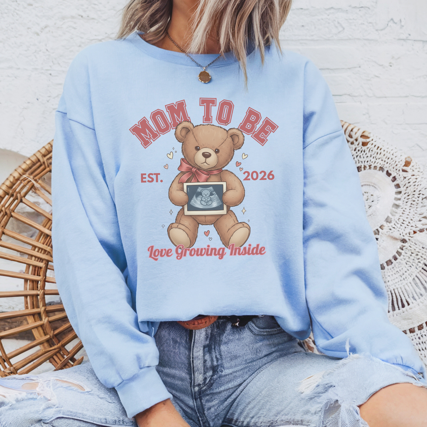 Light blue sweatshirt with teddy bear graphic and text, worn by a person sitting on a woven chair.