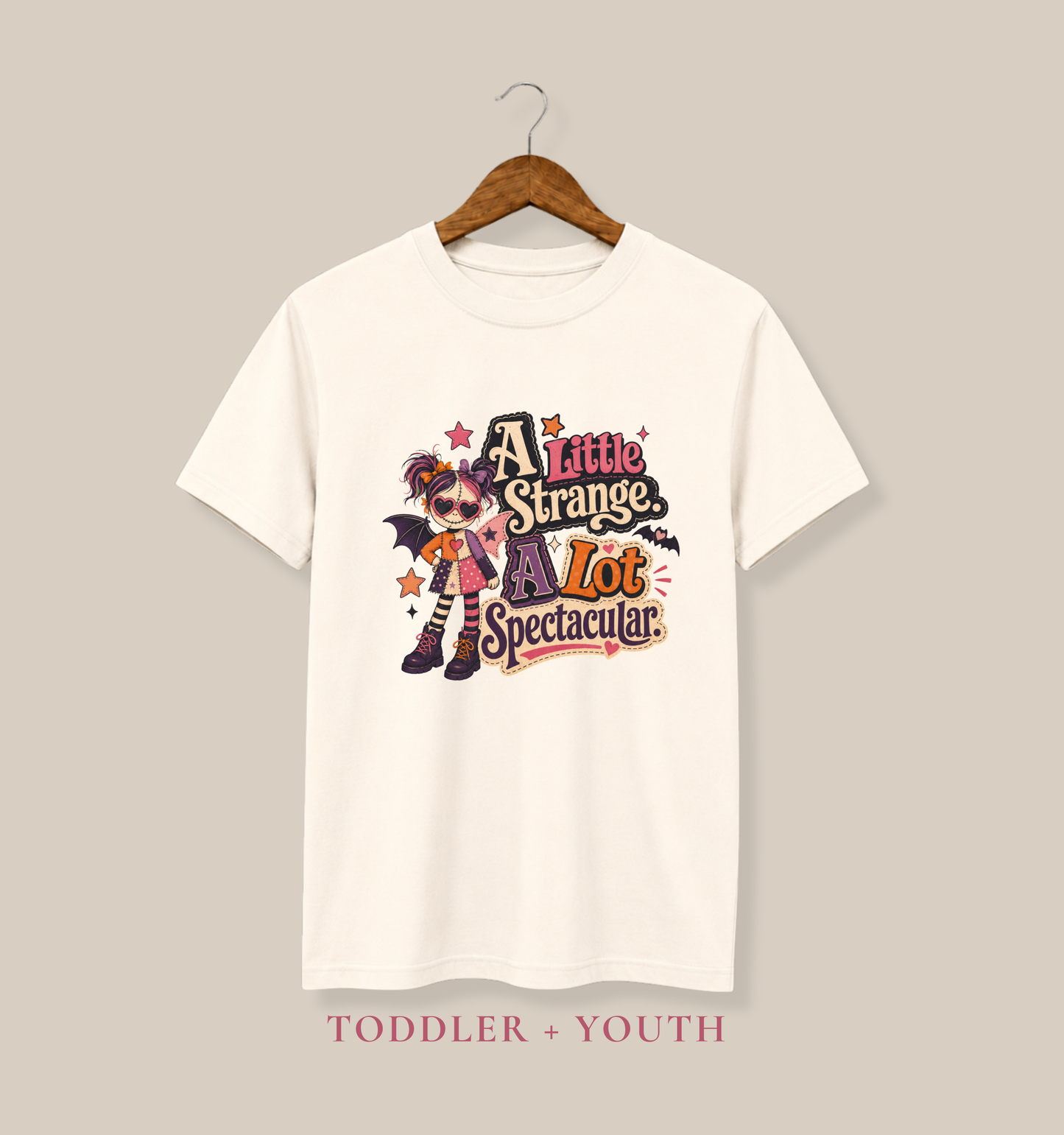 Natural youth T-shirt featuring a quirky spooky doll with colorful hearts, stars, bat wings, and the phrase “A Little Strange, A Lot Spectacular.”