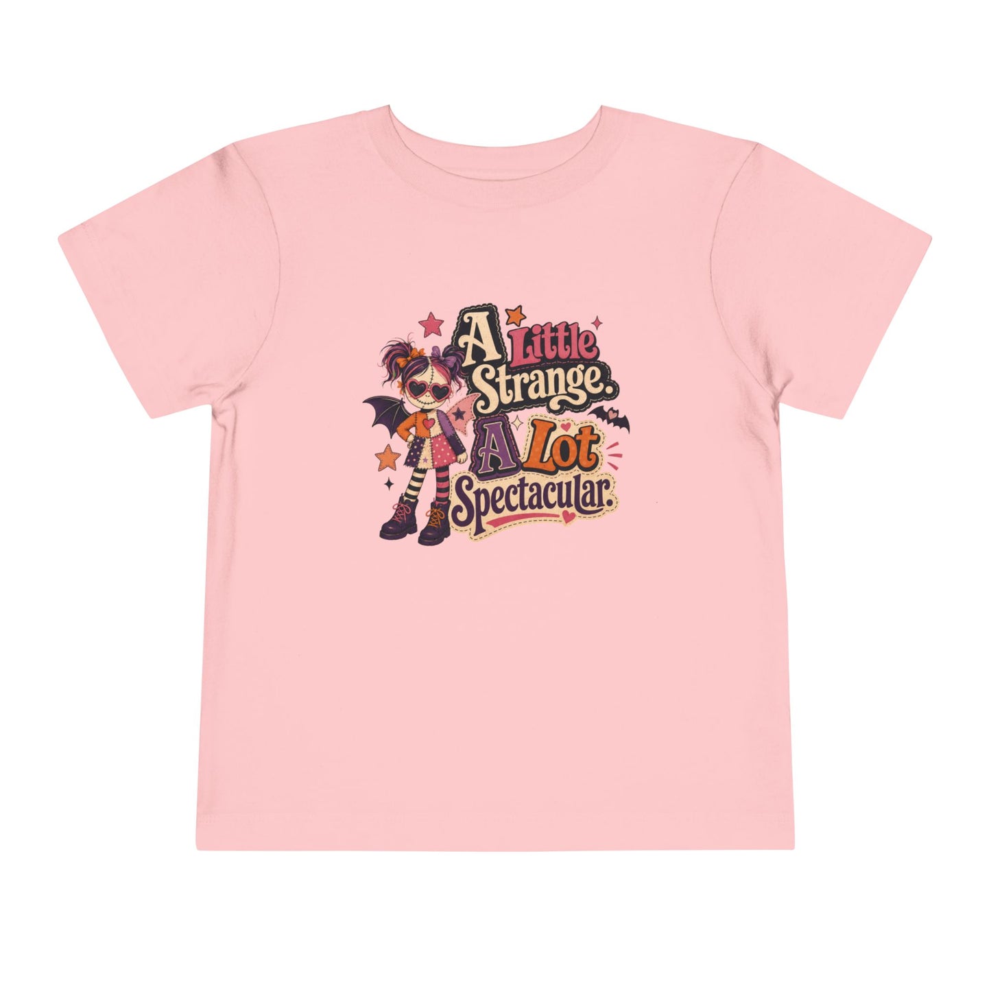 Pink toddler T-shirt featuring a quirky patchwork doll, hearts, stars, bat wings, and the phrase “A Little Strange, A Lot Spectacular.”