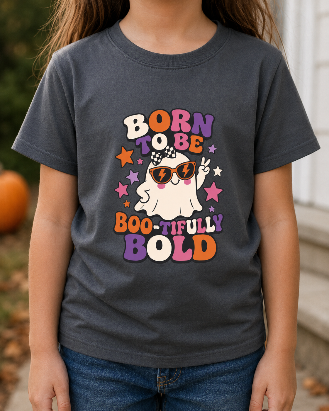A child wearing a dark charcoal gray graphic t-shirt with a colorful Halloween-themed design that reads 'BORN TO BE BOO-TIFULY BOLD' around a cartoon ghost wearing sunglasses. The background is a softly blurred outdoor setting with a pumpkin and a white door.