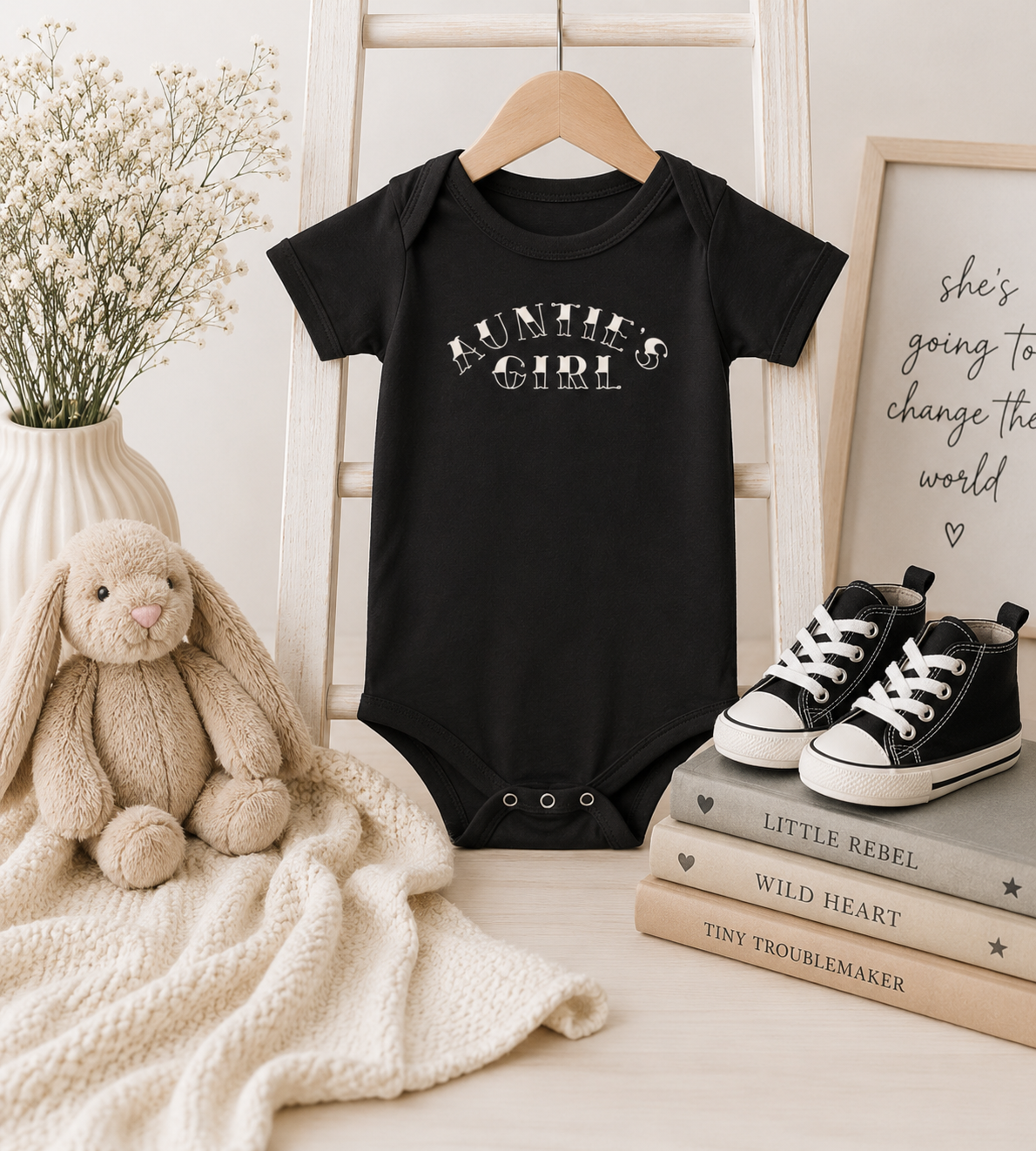 Black Bella + Canvas 100B infant one piece featuring a cream “Auntie’s Girl” graphic displayed in a cozy nursery mockup with neutral decor, stuffed bunny, baby shoes, and soft beige accents.