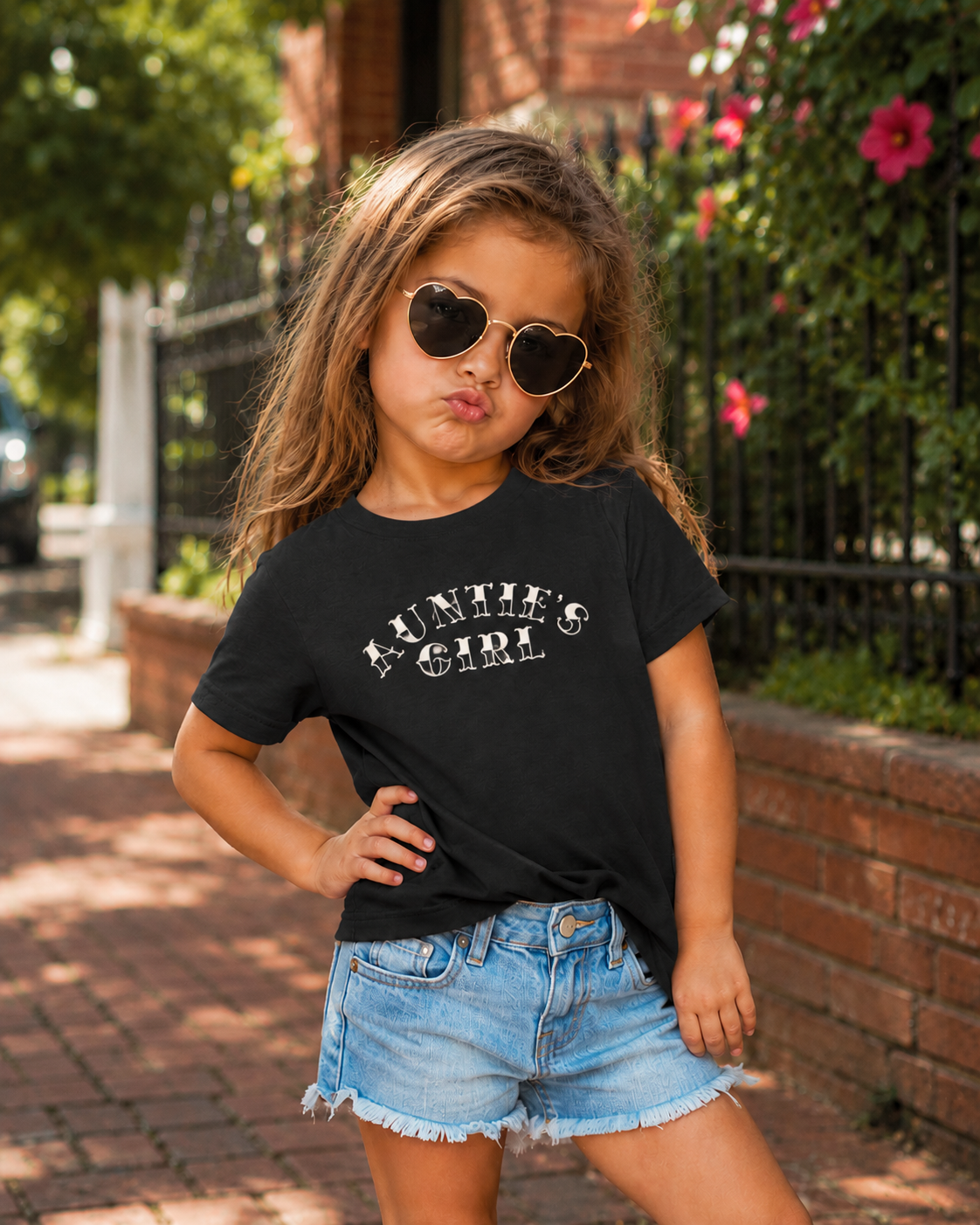 Young girl wearing a black Bella + Canvas 3001T toddler t-shirt with white “Auntie’s Girl” graphic lettering, styled with denim shorts and sunglasses in a trendy outdoor lifestyle mockup.