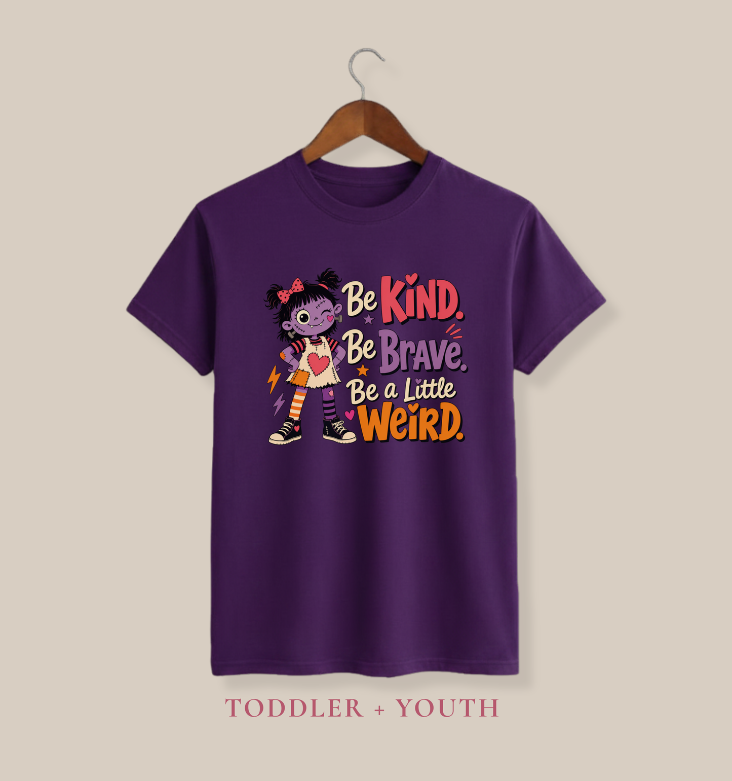 Team purple youth T-shirt featuring a quirky stitched doll, colorful accents, and the phrase “Be Kind, Be Brave, Be a Little Weird.”