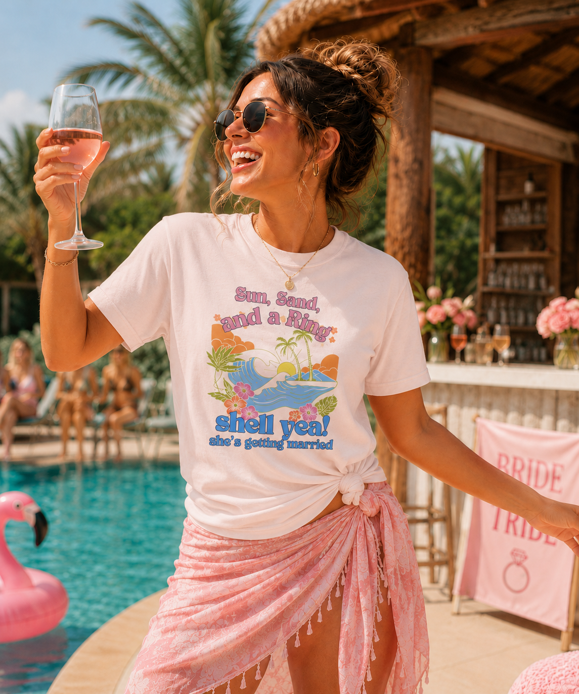 Woman wearing a Bella + Canvas 3001 t-shirt in soft pink with a tropical beach bachelorette design featuring palm trees, ocean waves, flowers, and “Sun, Sand, and a Ring, Shell Yeah! She’s Getting Married” wording at a poolside bridal party.