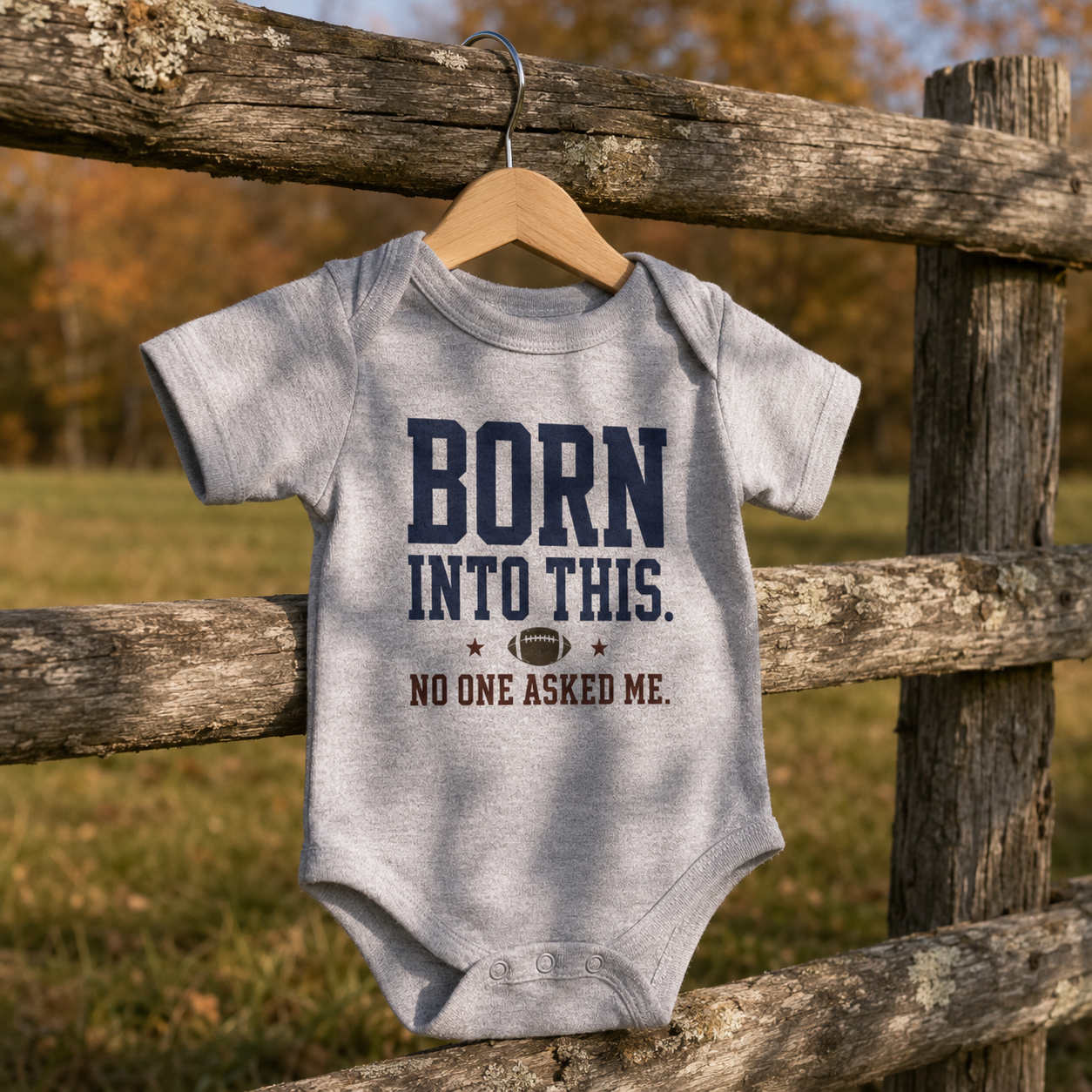 Bella + Canvas 100B infant one piece in Athletic Heather Grey featuring the phrase "Born Into This. No One Asked Me." with a football graphic. Soft football-themed baby bodysuit displayed on a rustic wooden fence with a fall background, perfect for newborns, infants, baby's first football season, game days, tailgates, baby showers, and football-loving families.