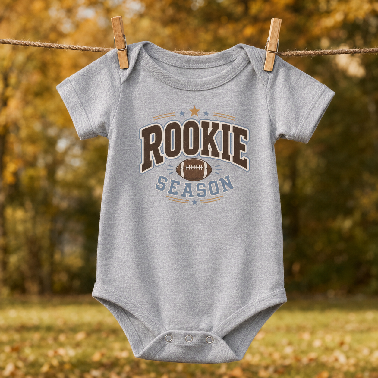 Bella + Canvas 100B infant one piece in Athletic Heather Grey featuring a football graphic and the words "Rookie Season." Soft football-themed baby bodysuit hanging on a clothesline outdoors with a fall background, perfect for newborns, infants, baby's first football season, game days, tailgates, baby showers, and sports-loving families.