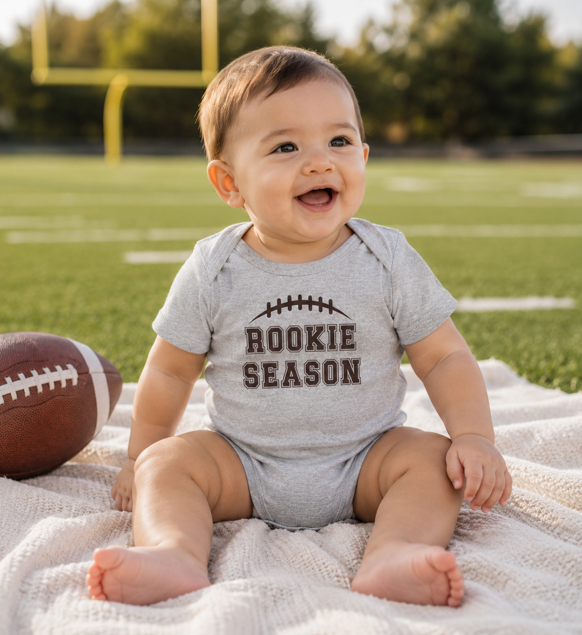 Smiling baby wearing a Bella + Canvas 100B athletic heather infant bodysuit featuring a brown football and "Rookie Season" design while sitting on a blanket at a football field. Perfect for football season, game day photos, baby showers, and newborn sports fans.