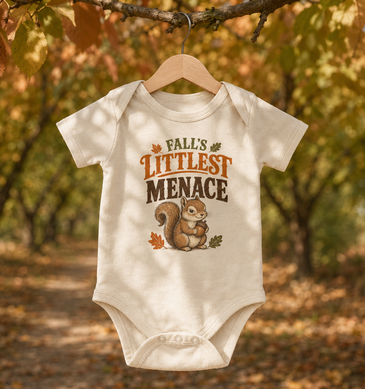 Natural Bella + Canvas 100B baby one piece hanging outdoors beneath autumn tree branches, featuring a cute squirrel holding an acorn with the phrase "Fall's Littlest Menace." Adorable fall baby bodysuit perfect for pumpkin patches, Halloween, autumn family photos, baby showers, and seasonal outings.