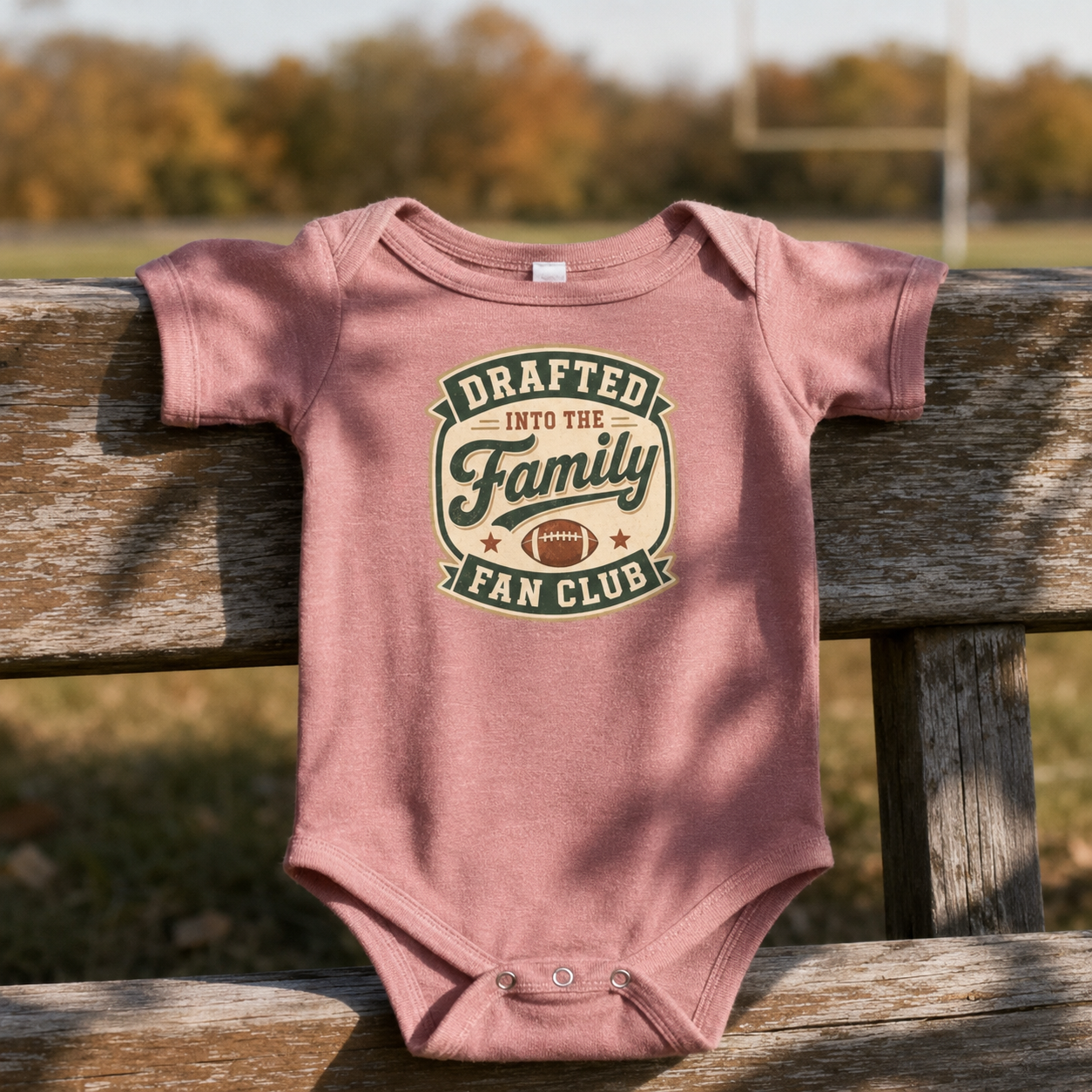Bella + Canvas 100B infant one piece in Heather Mauve featuring the phrase "Drafted Into the Family Fan Club" with a vintage football badge design. Soft football-themed baby bodysuit displayed on a rustic wooden fence at a football field, perfect for newborns, infants, baby's first football season, game days, tailgates, baby showers, football-loving families, and new sports fans.