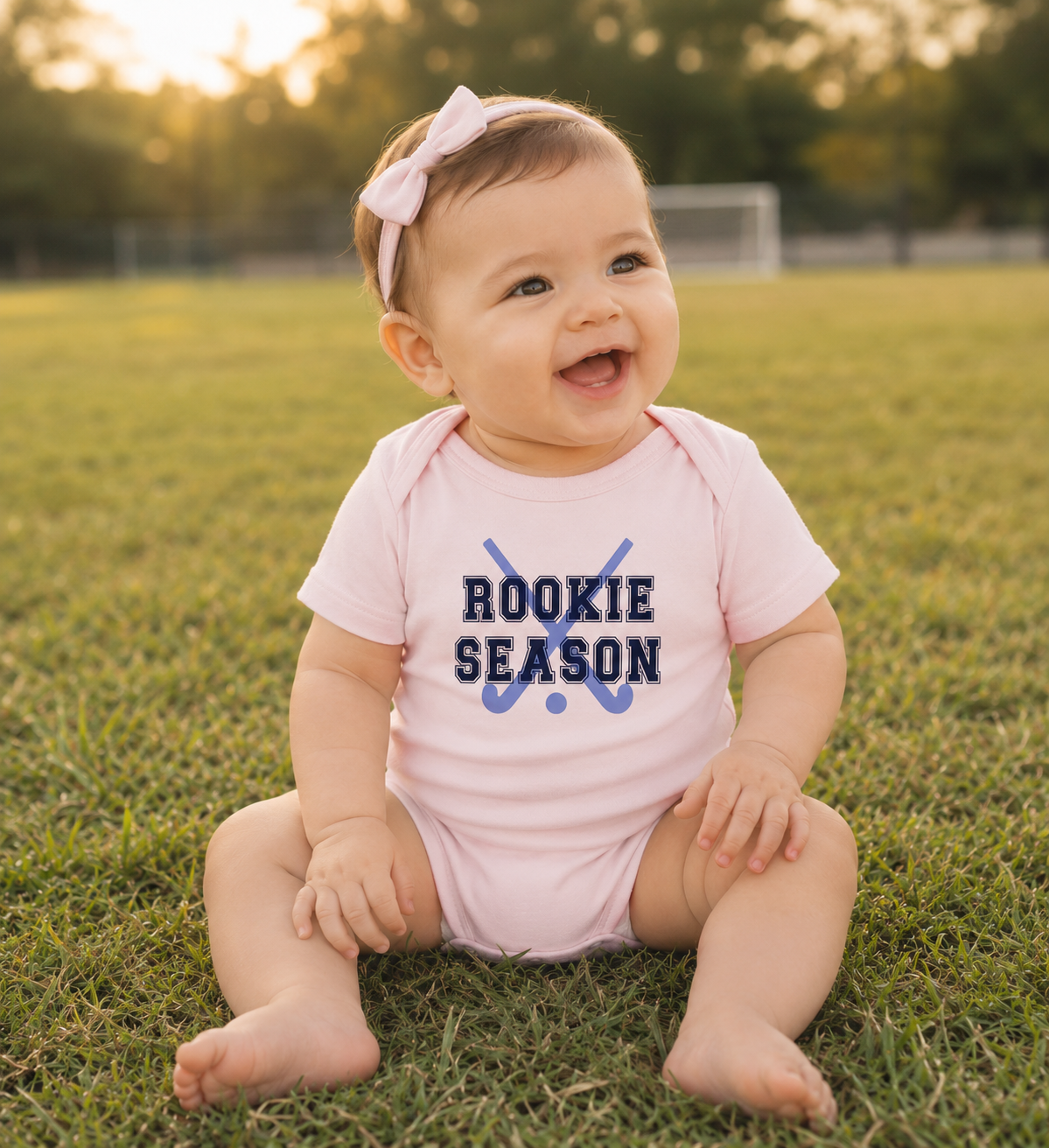 Bella + Canvas 100B pink infant bodysuit featuring crossed field hockey sticks and "Rookie Season" text. Adorable field hockey baby outfit for little girls, field hockey families, baby showers, newborn photos, and baby's first field hockey season.
