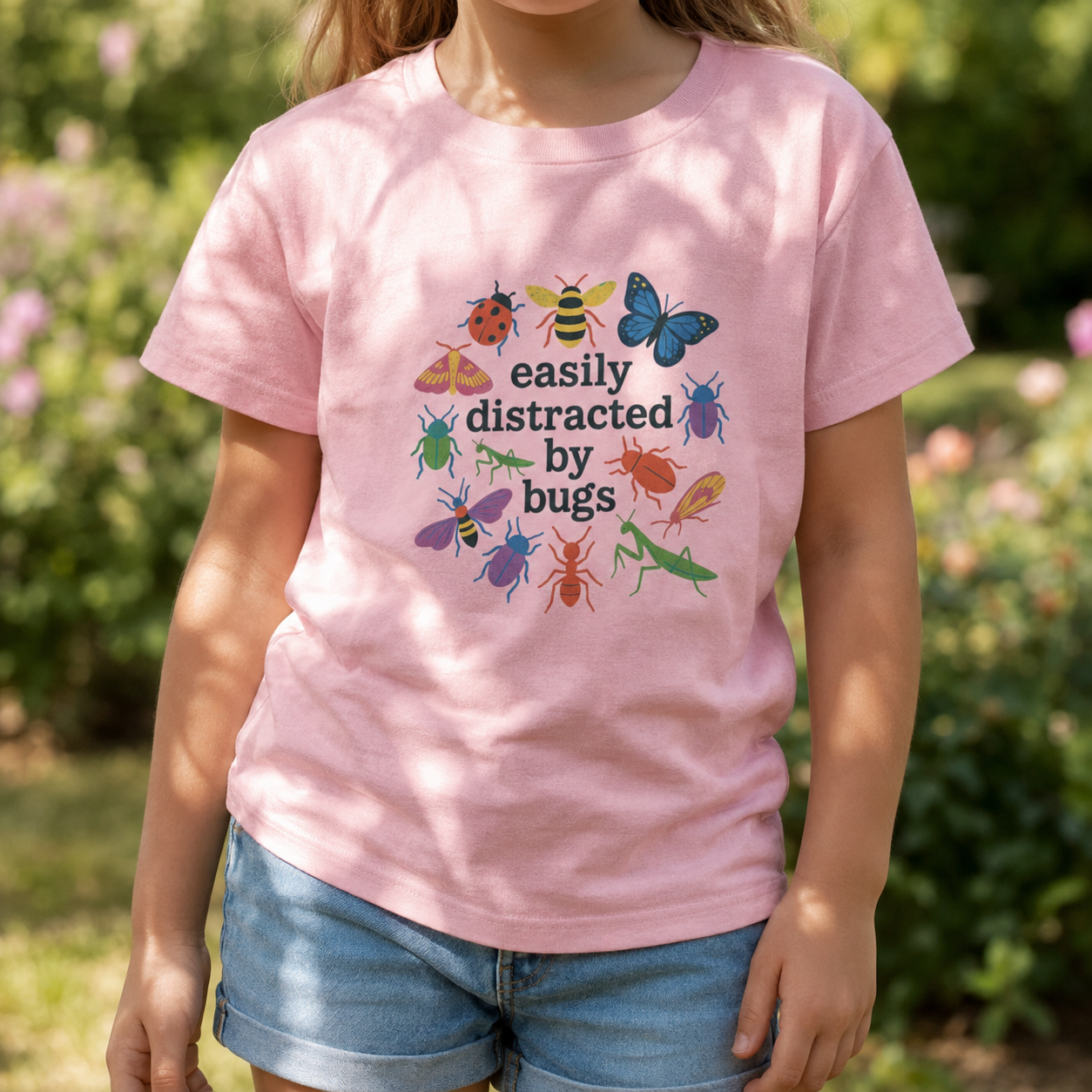 Bella + Canvas 3001Y youth t-shirt in Pink featuring the playful phrase "Easily Distracted by Bugs" surrounded by colorful insects including butterflies, bees, ladybugs, beetles, ants, and praying mantises. Fun nature-themed graphic tee for bug lovers, young explorers, homeschool, outdoor adventures, and everyday wear.