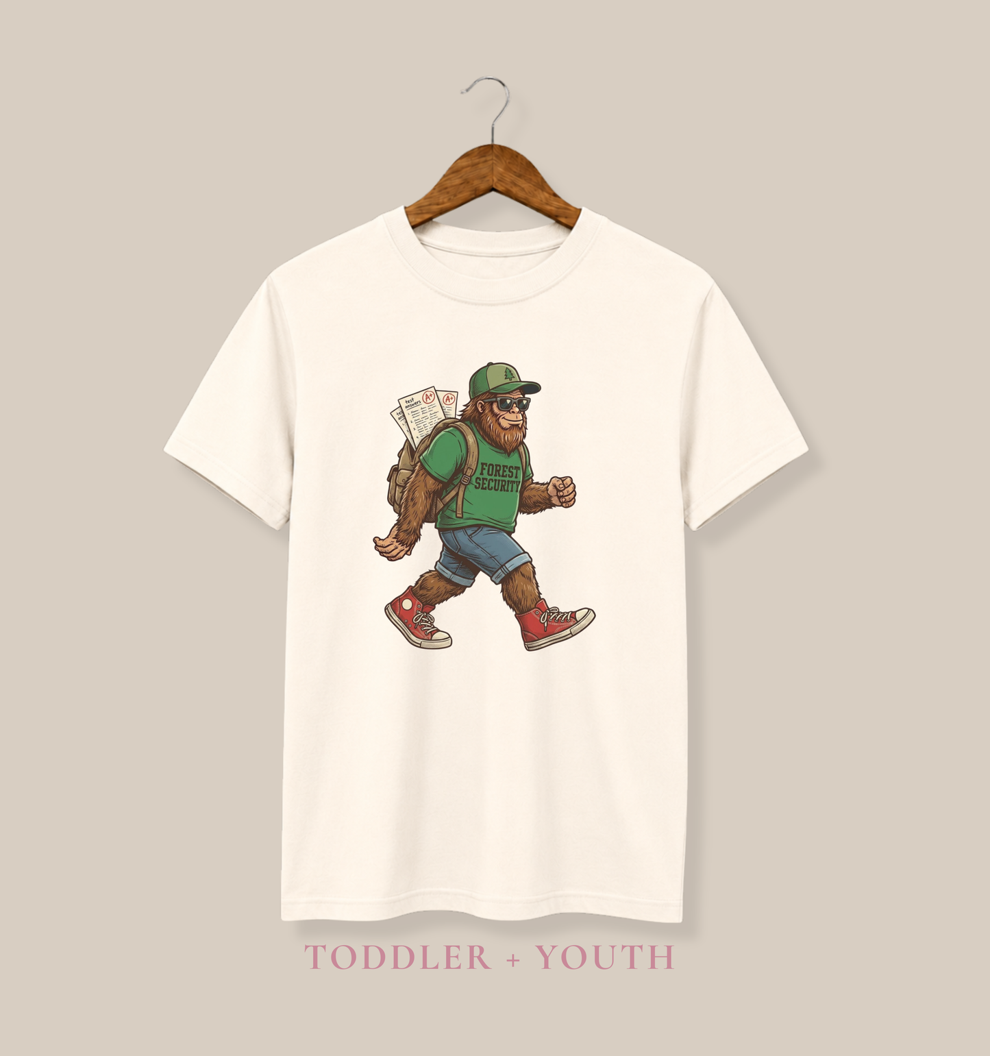 Bella + Canvas 3001Y youth t-shirt in natural featuring a Bigfoot student graphic wearing sunglasses, a backpack, and a green “Forest Security” shirt while carrying graded school papers.