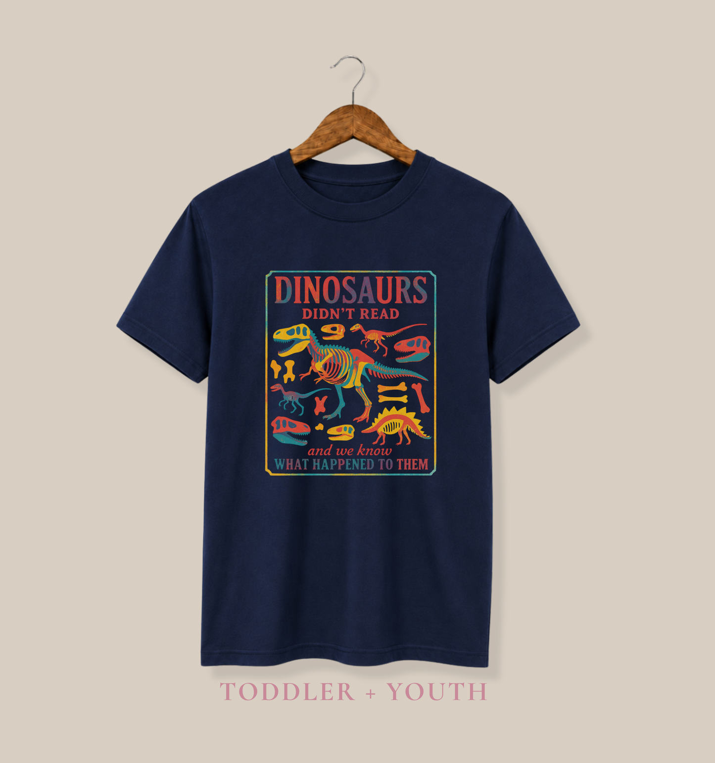 Bella + Canvas 3001Y youth t-shirt in navy featuring a colorful dinosaur skeleton graphic with the humorous quote “Dinosaurs Didn’t Read and We Know What Happened to Them.” Fun reading-themed shirt for kids displayed on a navy tee hanging from a wooden hanger.