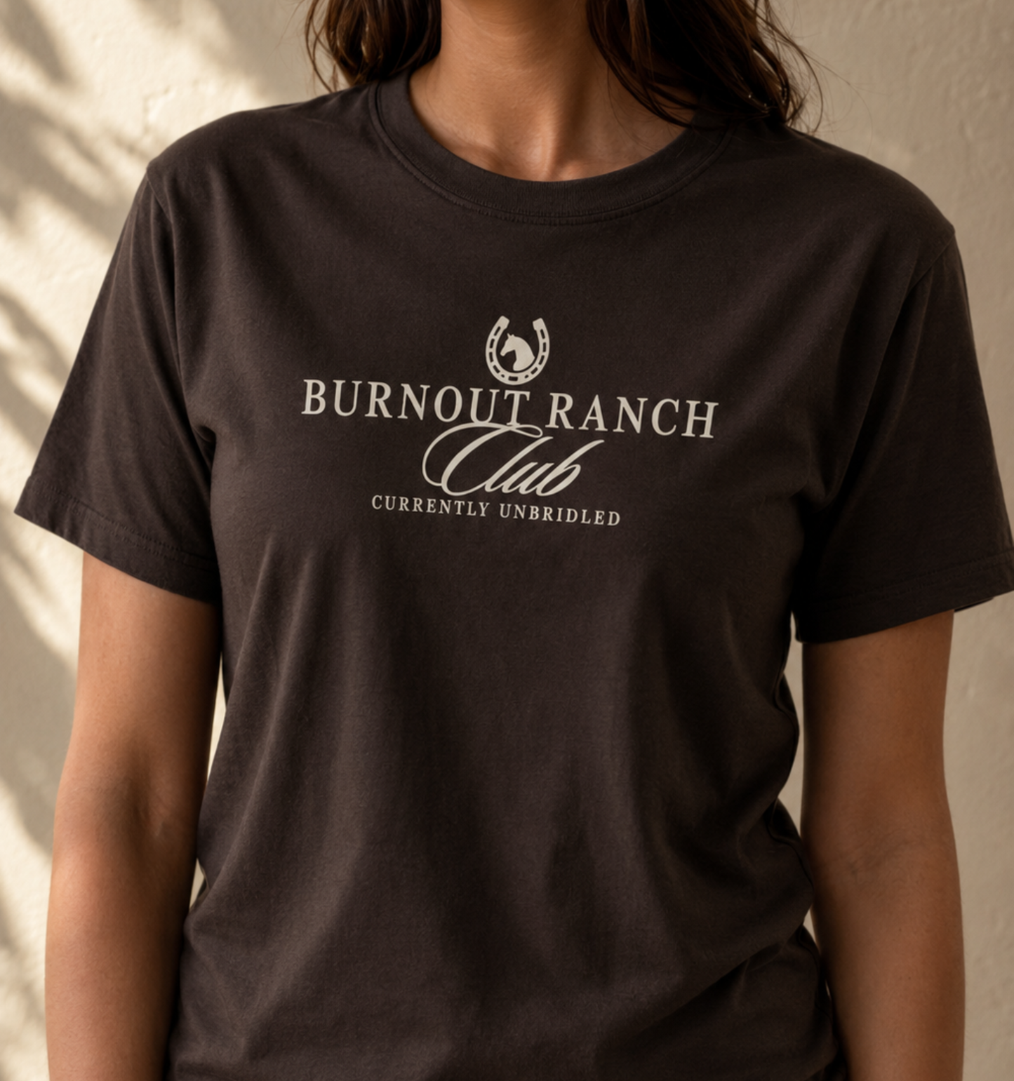 Bella + Canvas 3001 unisex t-shirt in Brown featuring the "Burnout Ranch Club" graphic with a horseshoe and horse silhouette above the phrase "Currently Unbridled." A western-inspired graphic tee perfect for horse lovers, rodeo fans, equestrians, ranch life enthusiasts, and casual everyday wear.