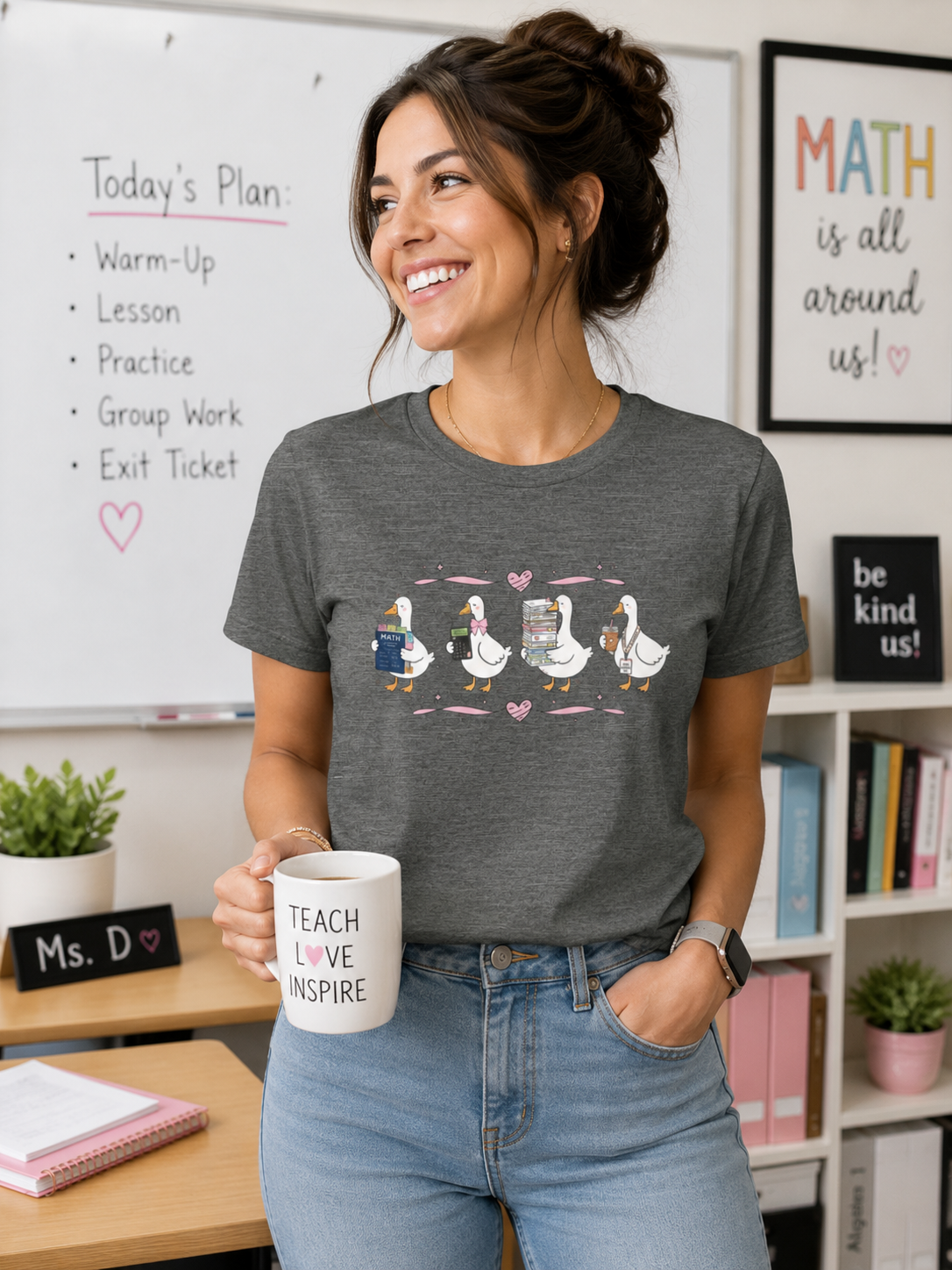 Female teacher wearing a Bella + Canvas 3001 t-shirt in Deep Heather featuring a math teacher goose design. The illustration shows four white geese holding a math textbook, calculator, stack of papers, and iced coffee with a teacher lanyard. Pink bows and heart accents add a coquette touch. Styled in a classroom setting, this fun math teacher shirt makes a great back-to-school outfit, teacher appreciation gift, or everyday educator tee.