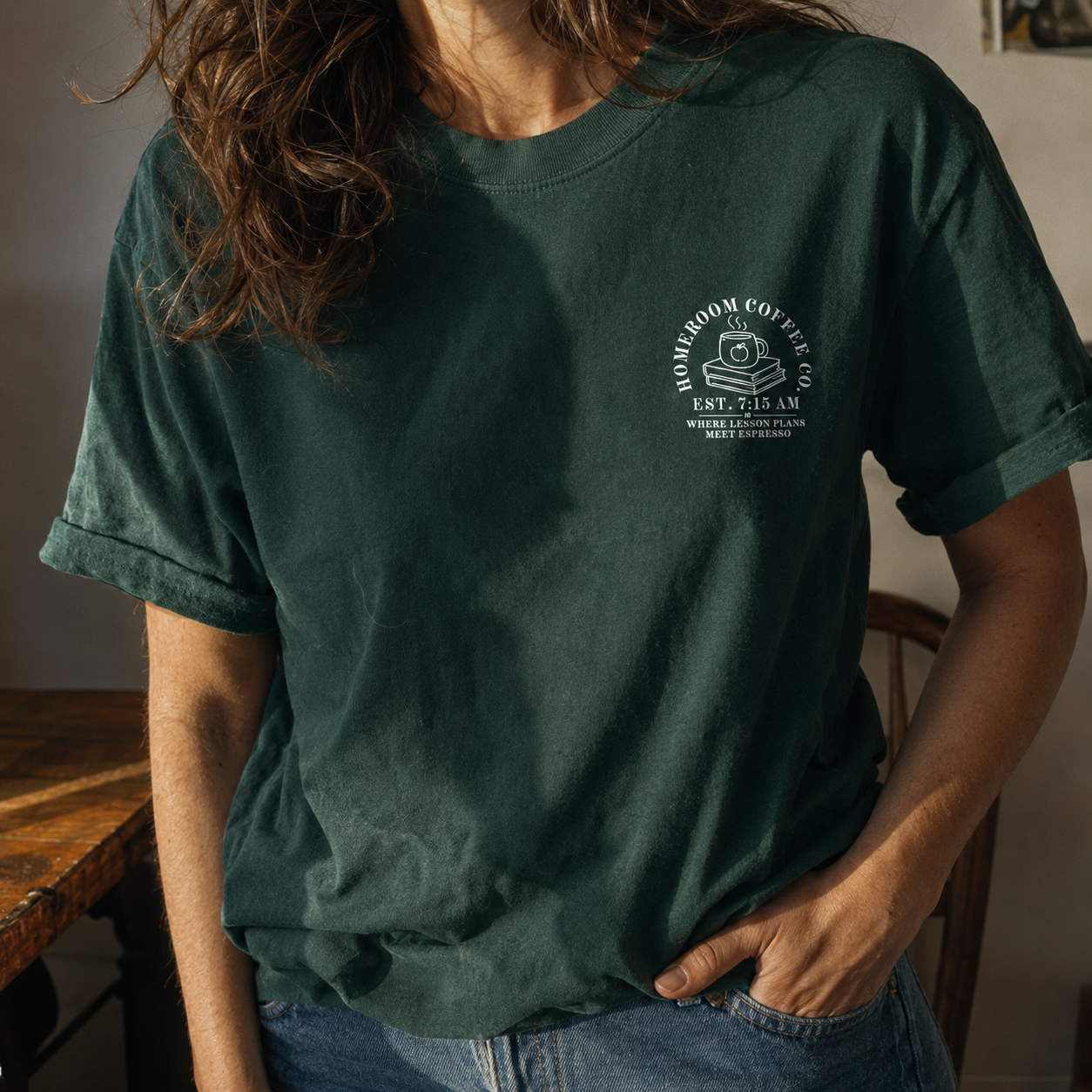 Female model wearing a Bella + Canvas 3001 t-shirt in Forest featuring a minimalist Homeroom Coffee Co. pocket design with a steaming coffee mug on stacked books and the phrase "Where Lesson Plans Meet Espresso." Styled with rolled sleeves in a warm, cozy setting. Perfect for teachers, educators, coffee lovers, and back-to-school outfits.