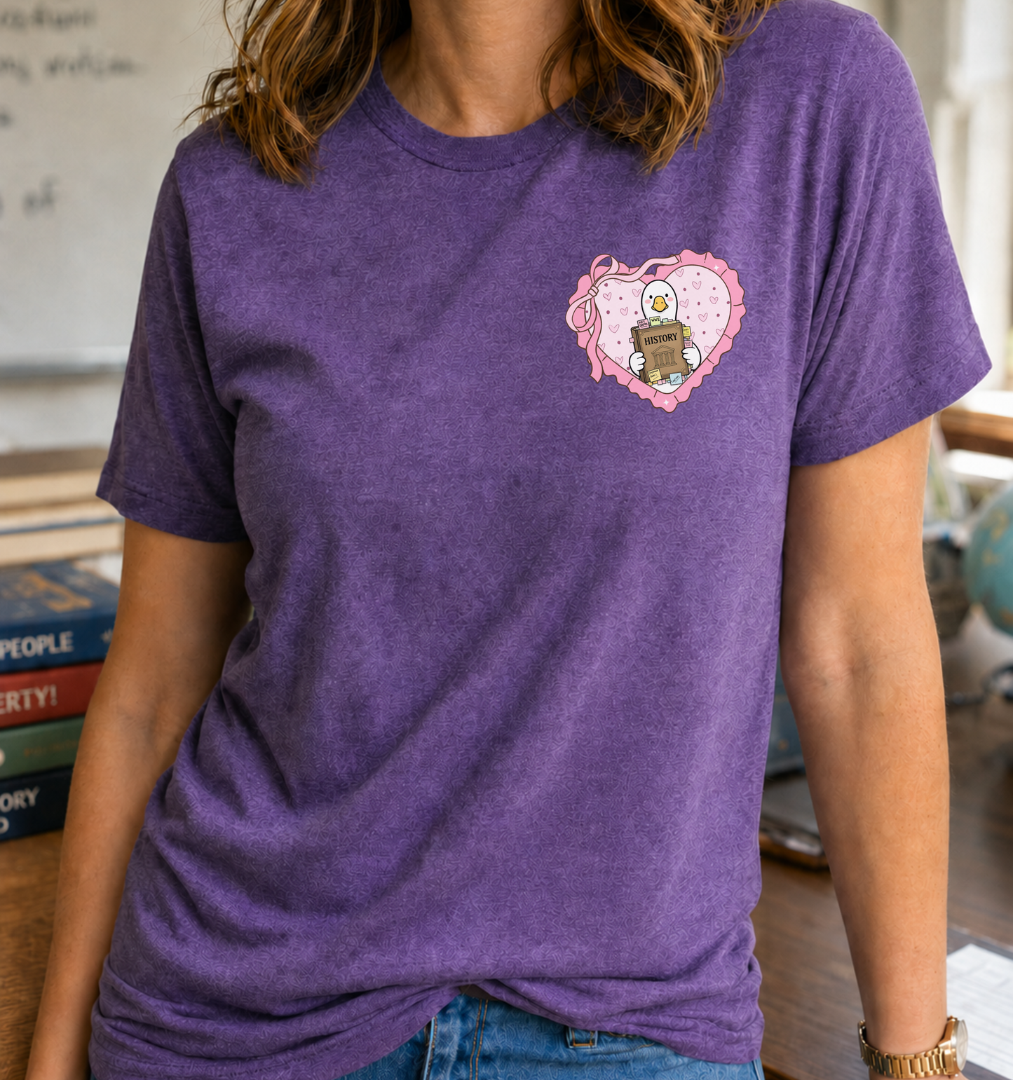 Bella + Canvas 3001 unisex t-shirt in Heather Team Purple featuring a heart-shaped pocket graphic with a cute goose holding a history textbook, surrounded by pink hearts and a ribbon. A fun history teacher shirt perfect for educators, social studies teachers, classroom wear, and history lovers.