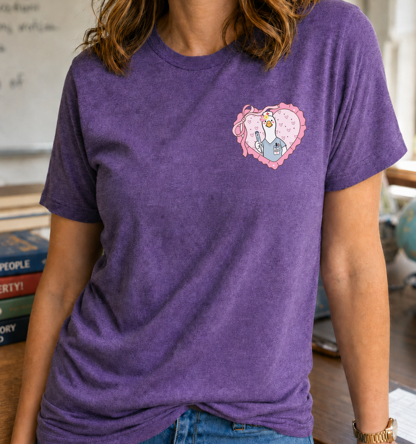 Bella + Canvas 3001 unisex t-shirt in Heather Team Purple featuring a cute goose dressed as a school nurse inside a pink heart pocket graphic, wearing scrubs, holding a thermometer, and a school nurse badge. A fun school nurse shirt perfect for nurses, healthcare staff, back-to-school, Nurse Appreciation Week, and everyday wear.