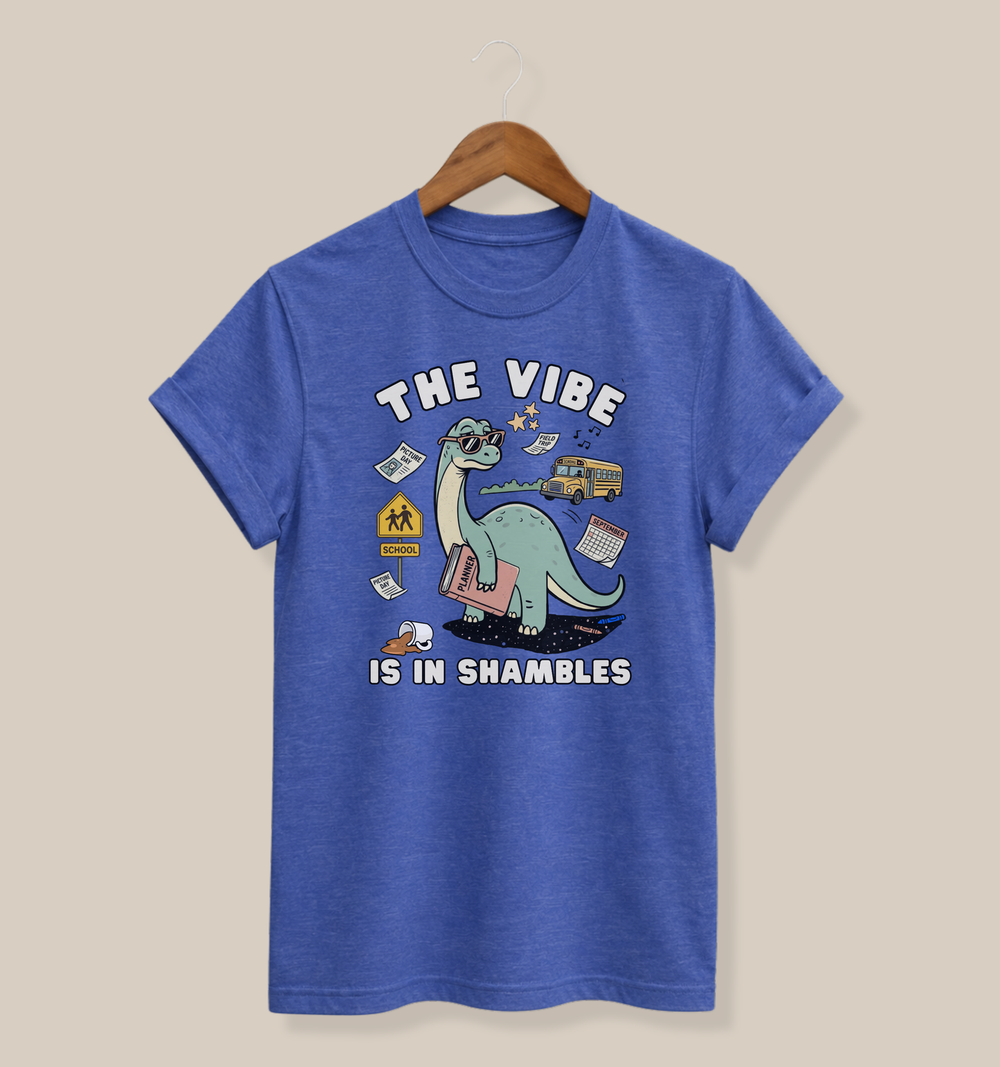 Bella + Canvas 3001 heather true royal blue t-shirt featuring a funny teacher dinosaur design with the phrase "The Vibe Is In Shambles." The graphic includes a dinosaur holding a planner surrounded by a school bus, classroom supplies, a spilled coffee cup, a calendar, and back-to-school icons. Perfect for teachers, educators, school staff, elementary teachers, and anyone navigating the chaos of the school year with humor.