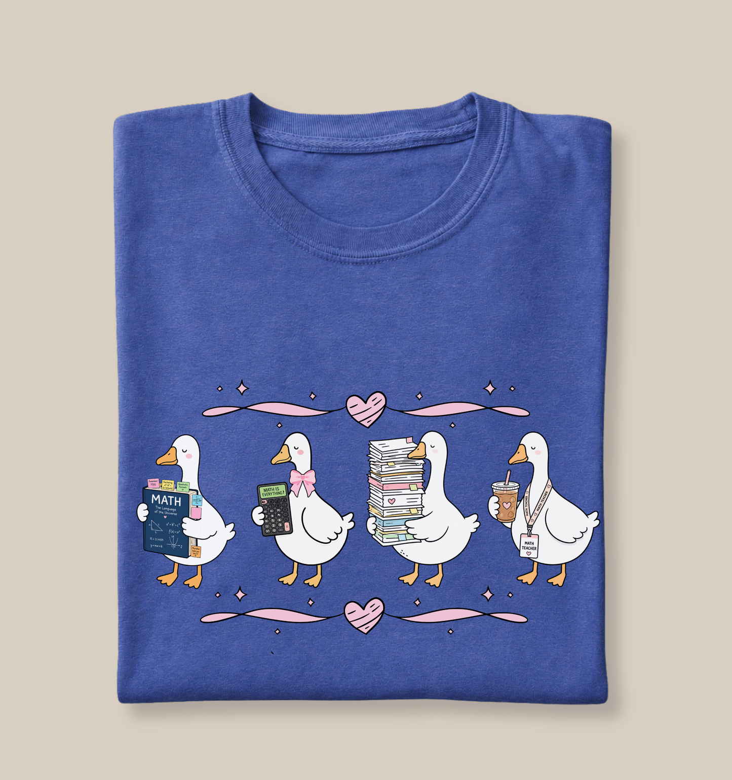 Folded Bella + Canvas 3001 t-shirt in Heather True Royal featuring a whimsical math teacher goose design. The illustration shows four white geese carrying a math textbook, calculator, stack of papers, and iced coffee with a teacher badge. Pink bows, hearts, and coquette-inspired accents complete the design. Perfect for math teachers, educators, back-to-school outfits, teacher appreciation gifts, and classroom wear.