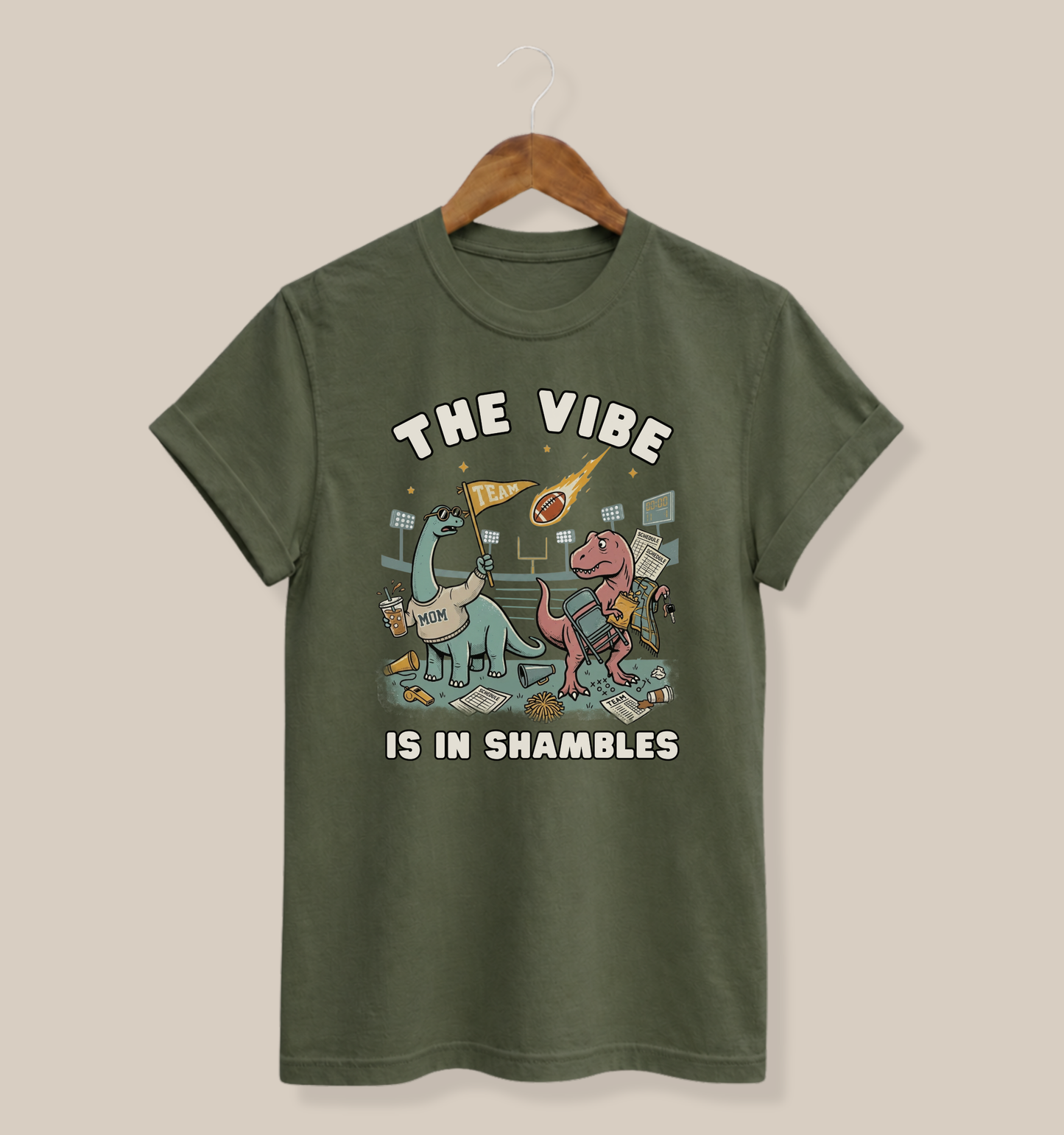 Bella + Canvas 3001 military green t-shirt featuring two overwhelmed dinosaurs at a sports field surrounded by a folding chair, snacks, schedule papers, coffee, and team gear. Funny sports mom graphic tee with "The Vibe Is In Shambles" design, perfect for football moms, busy sports parents, game day outfits, and sports season gifts.