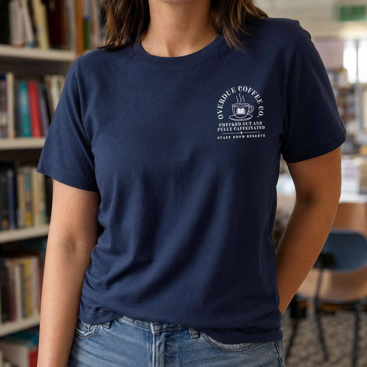Bella + Canvas 3001 unisex t-shirt in Navy worn in a library setting, featuring a white Overdue Coffee Co. pocket-style graphic with a steaming coffee cup and open book illustration. The design reads "Checked Out and Fully Caffeinated" and "Staff Room Reserve," making it a perfect graphic tee for librarians, library staff, teachers, book lovers, and coffee enthusiasts.