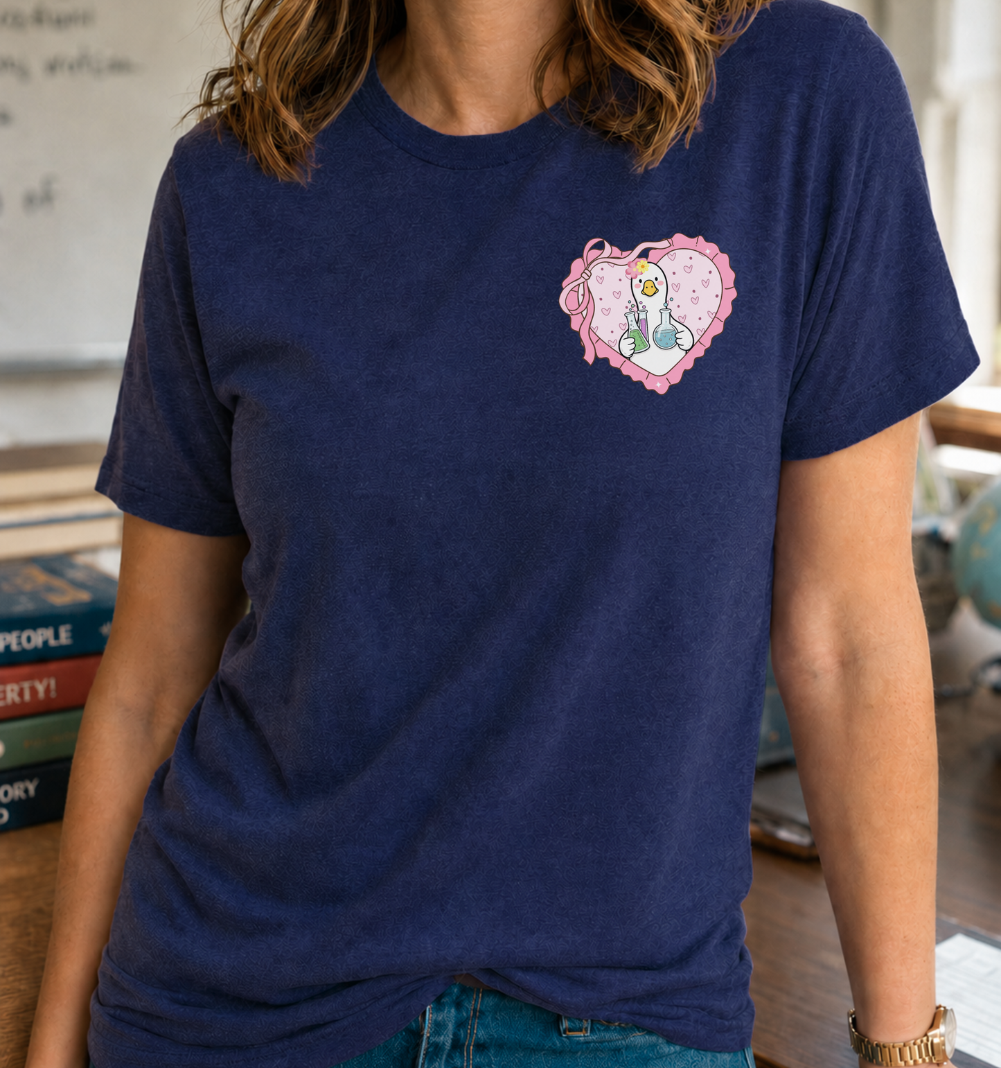 Bella + Canvas 3001 unisex t-shirt in Navy featuring a cute goose holding colorful laboratory beakers inside a pink heart-shaped pocket graphic with ribbon and heart details. A fun science teacher shirt perfect for chemistry teachers, STEM educators, science classrooms, back-to-school, and Teacher Appreciation gifts.