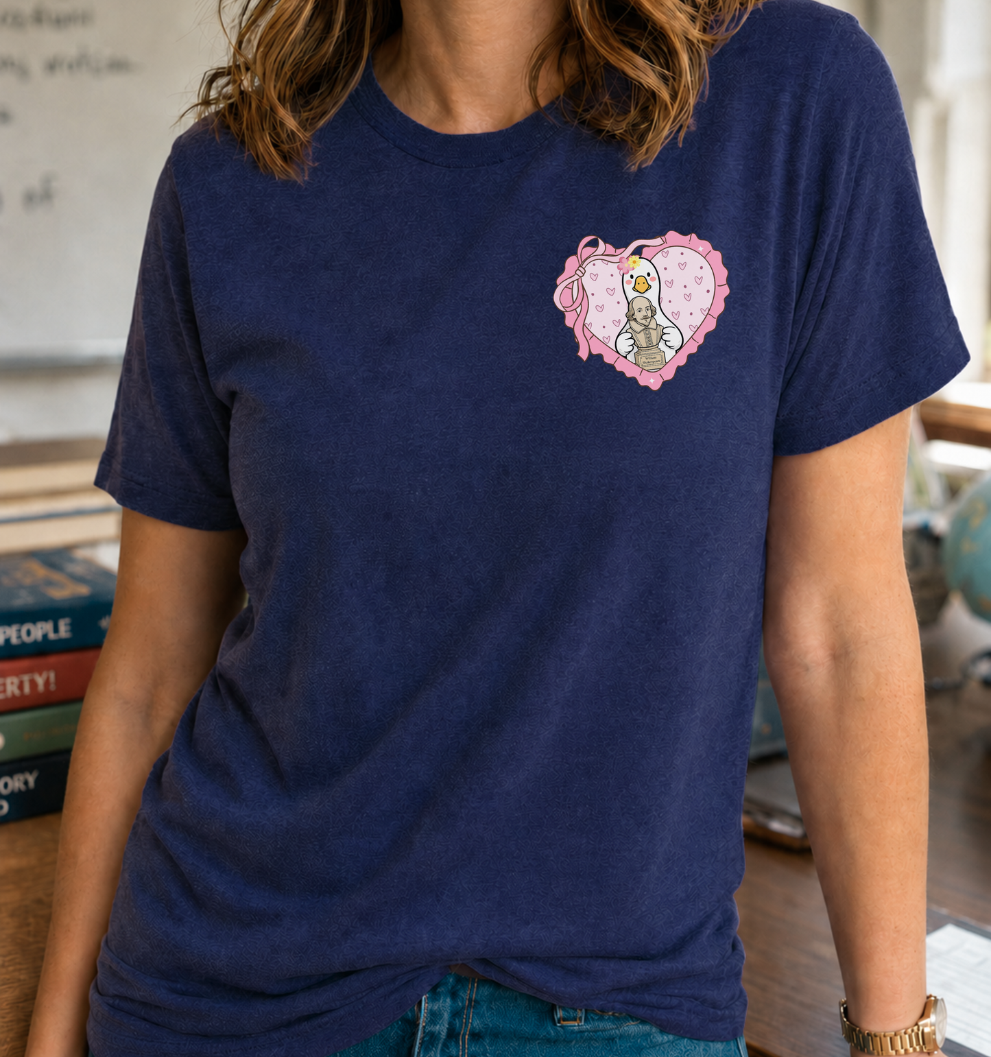 Bella + Canvas 3001 unisex t-shirt in Navy featuring a heart-shaped pocket graphic with a duck holding a Shakespeare bust, surrounded by pink hearts and a bow. A cute literary-inspired teacher shirt perfect for English teachers, literature lovers, Valentine's Day, and classroom wear.