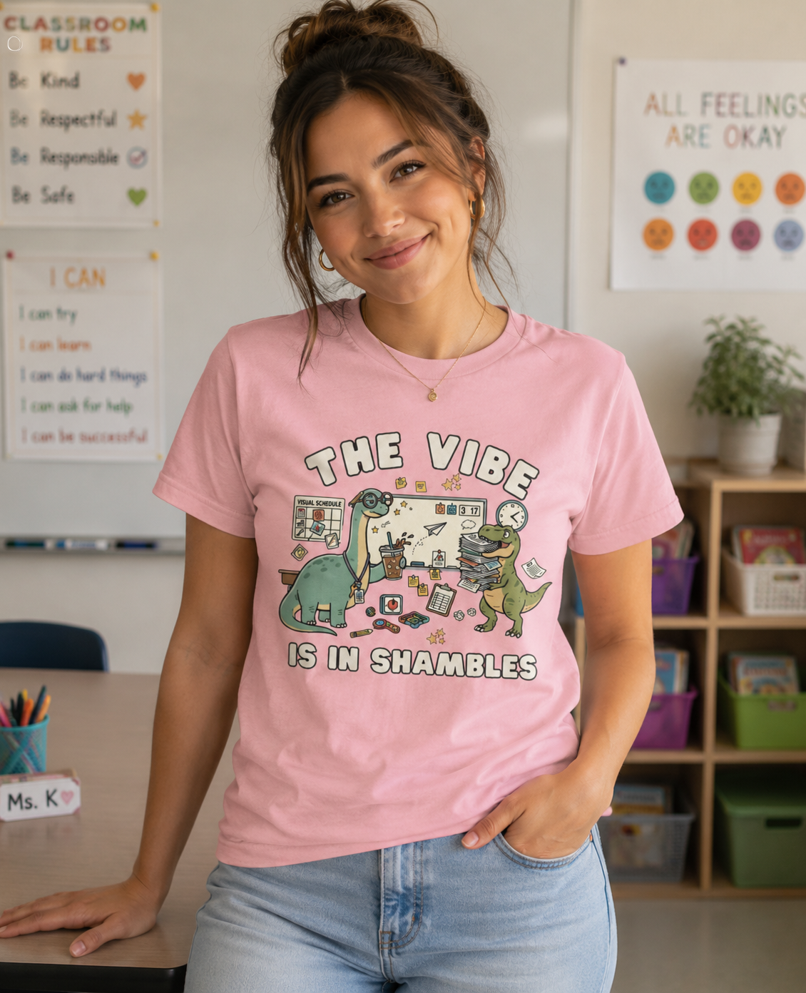 Young woman wearing a Bella + Canvas 3001 pink t-shirt in a classroom featuring a funny special education teacher dinosaur design with the phrase "The Vibe Is In Shambles." The graphic includes dinosaurs surrounded by visual schedules, classroom supplies, paperwork, fidget toys, and teaching tools. Perfect for special education teachers, IEP teachers, paraprofessionals, school staff, and educators who bring humor to busy classroom days.