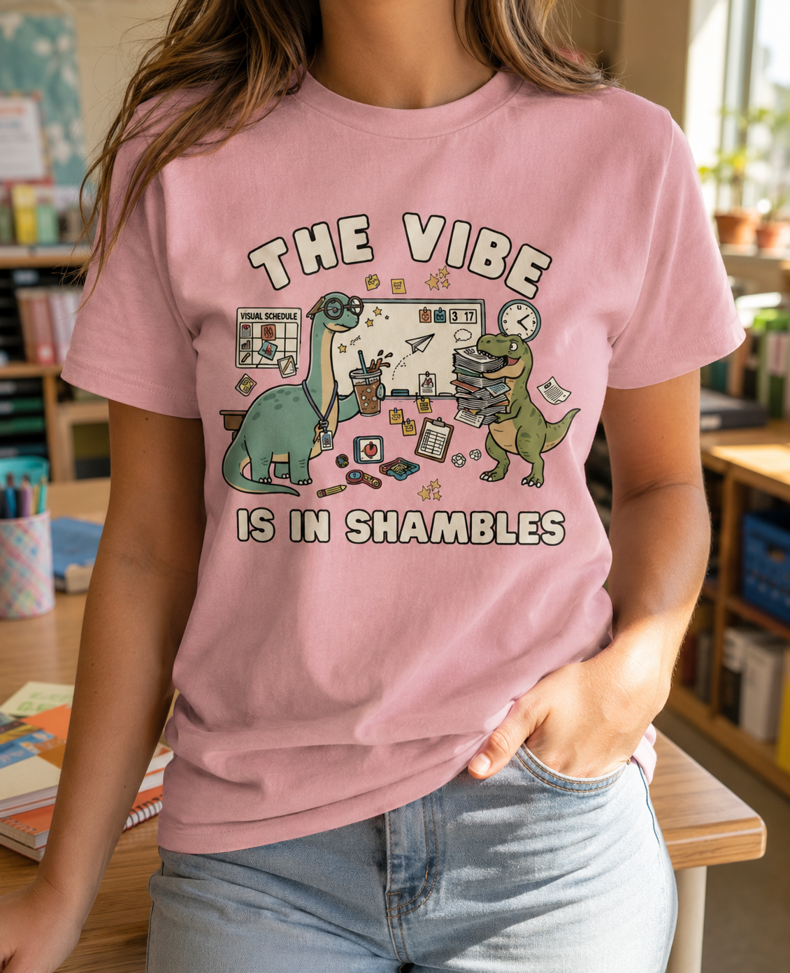 Bella + Canvas 3001 unisex t-shirt in Pink featuring the phrase "The Vibe Is in Shambles" with two adorable classroom dinosaurs, a visual schedule, books, coffee, and school supplies. A funny teacher shirt perfect for special education teachers, elementary educators, school staff, and back-to-school outfits.