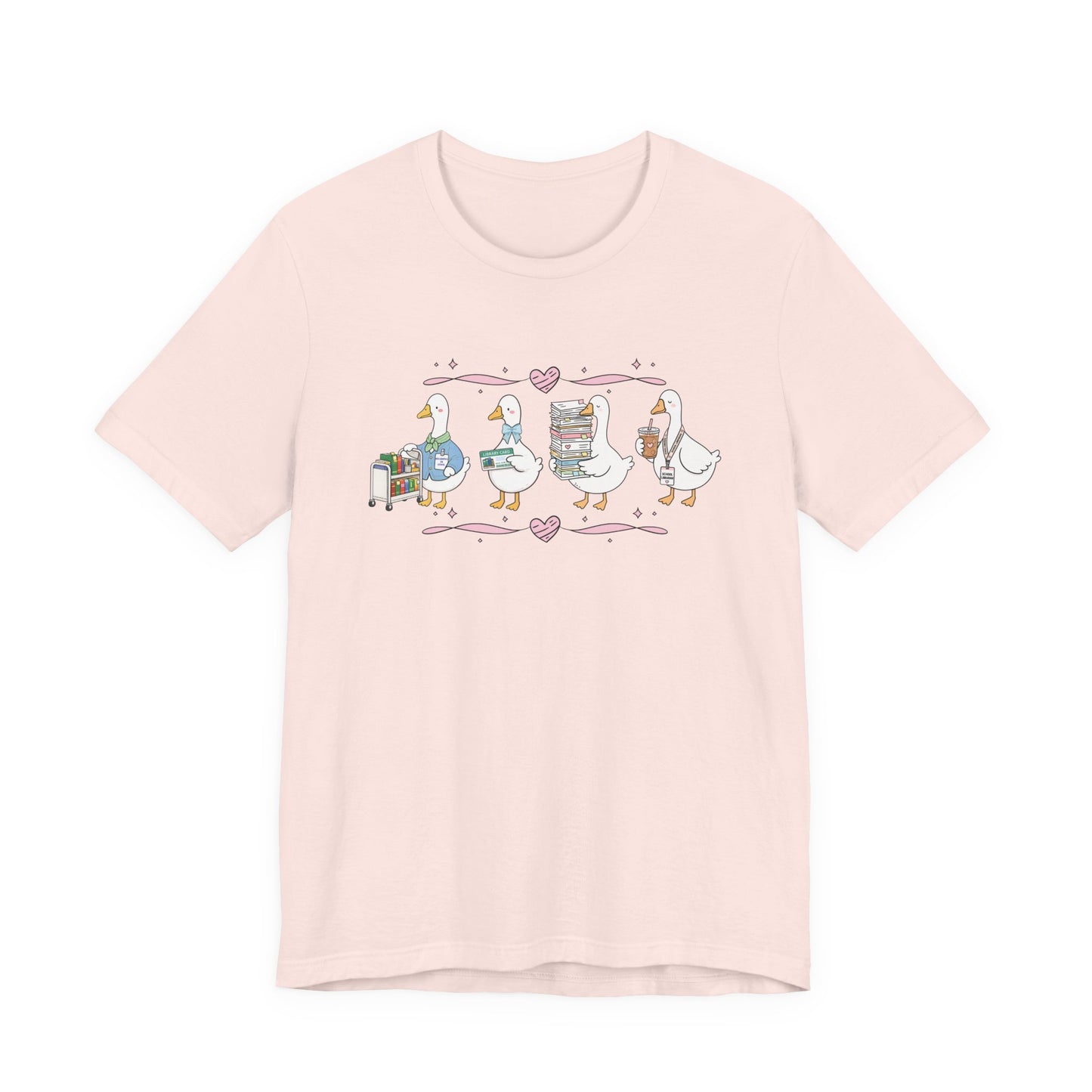 Bella + Canvas 3001 t-shirt in Soft Pink featuring a coquette librarian goose design. Four white geese display a library book cart, library card, stack of books, and iced coffee with a school librarian badge. Pink hearts and ribbon accents add a whimsical touch, making this cute librarian shirt perfect for school librarians, media specialists, library staff, book lovers, teacher appreciation gifts, and back-to-school outfits.