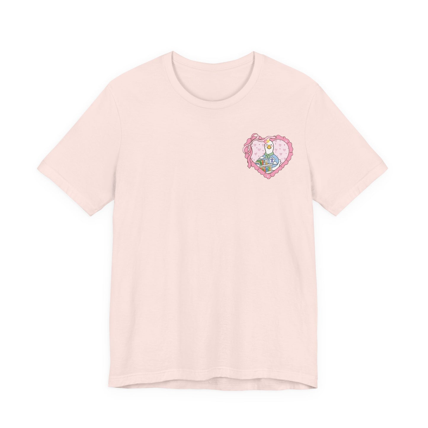 Bella + Canvas 3001 t-shirt in Soft Pink featuring a coquette school librarian goose pocket design. The artwork shows a white goose behind a colorful library book cart inside a pink heart with ribbon details. Cute librarian gift shirt for library staff and readers.