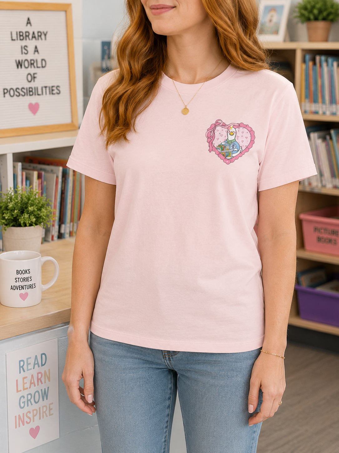 Female model wearing a Bella + Canvas 3001 t-shirt in Soft Pink featuring a coquette school librarian goose pocket design. The artwork shows a white goose behind a colorful book cart inside a pink heart with ribbon details. Photographed in a bright library setting with bookshelves and reading-themed decor. Ideal for school librarians, media specialists, library staff, and book lovers.