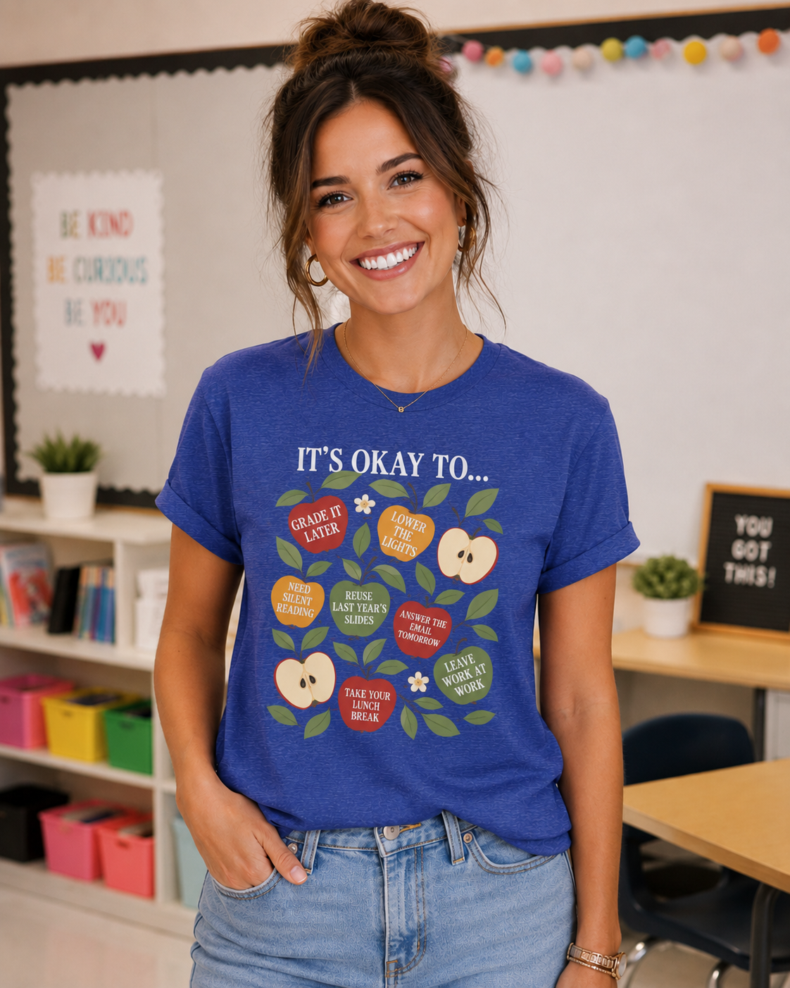 Woman wearing a Bella + Canvas 3001 t-shirt in true royal blue featuring colorful apple illustrations with teacher self care sayings including “Grade It Later,” “Lower the Lights,” and “Leave Work at Work” in a classroom setting.