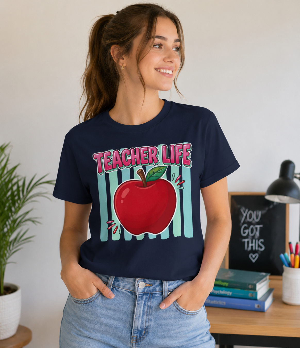 Young woman wearing a Bella + Canvas 3001 t-shirt in navy featuring a colorful Teacher Life graphic with a bright red apple and retro striped background. Casual teacher apparel shown in a classroom-inspired setting.