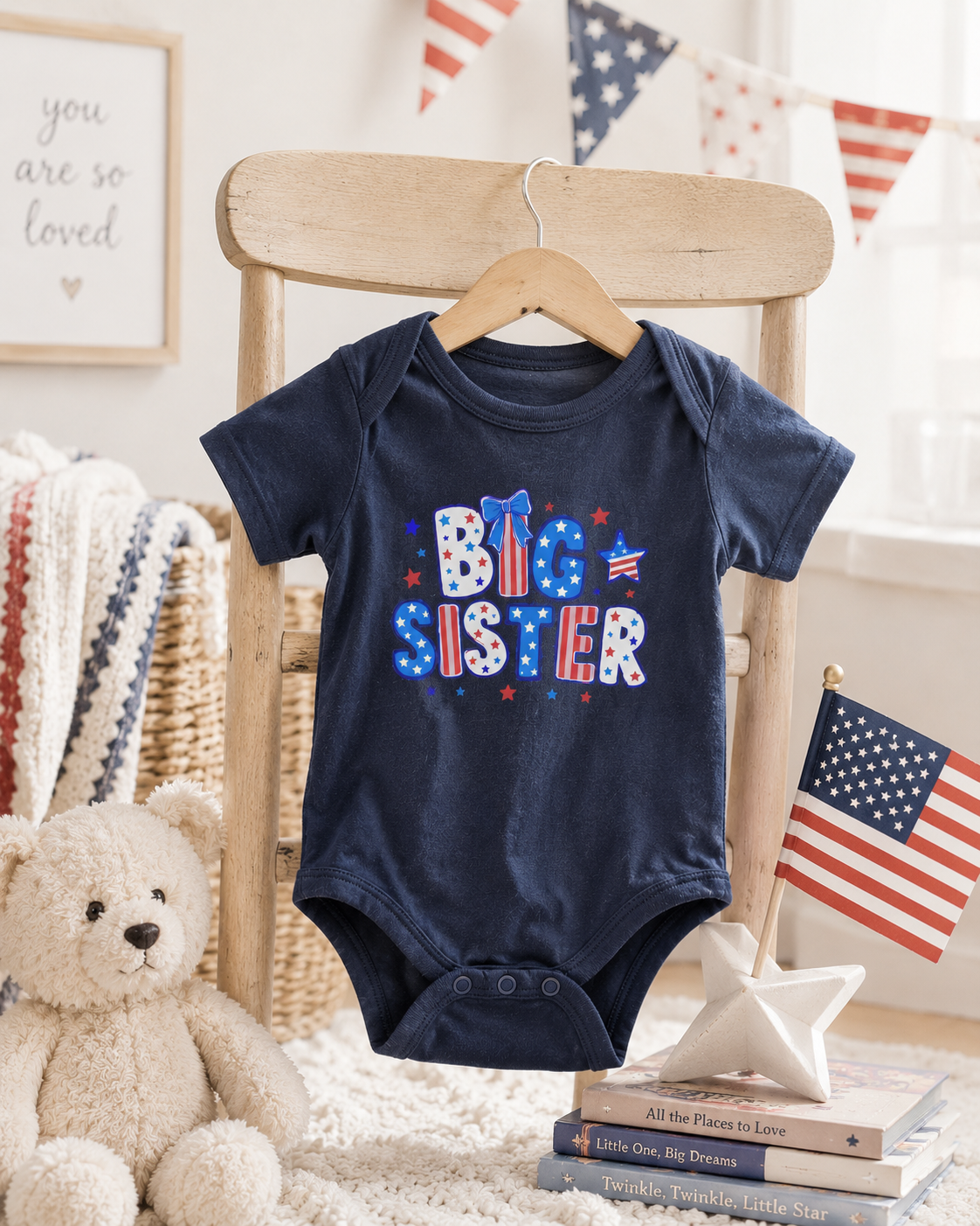Navy Bella + Canvas 100B infant one piece with patriotic “Big Sister” graphic displayed in a cozy nursery setting with American flag decor, teddy bear, and Fourth of July themed accents.