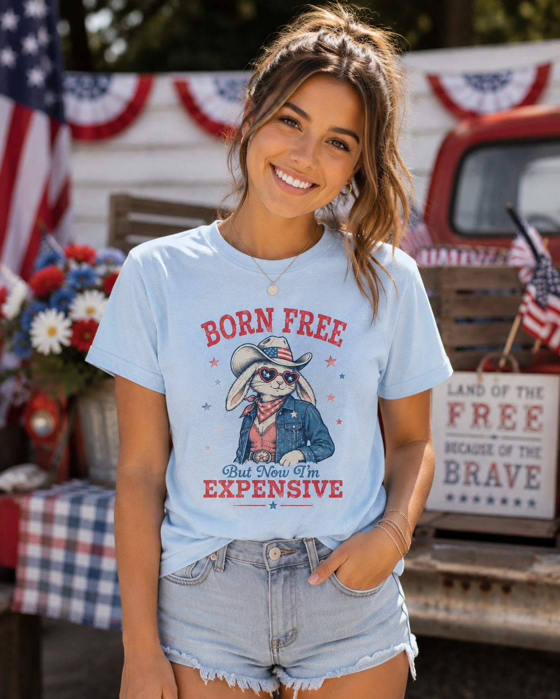 Adult Bella + Canvas baby blue t-shirt featuring a patriotic bunny cowgirl graphic with heart-shaped sunglasses, denim jacket, cowboy hat, and the phrase “Born Free But Now I’m Expensive” in vintage red and blue lettering.