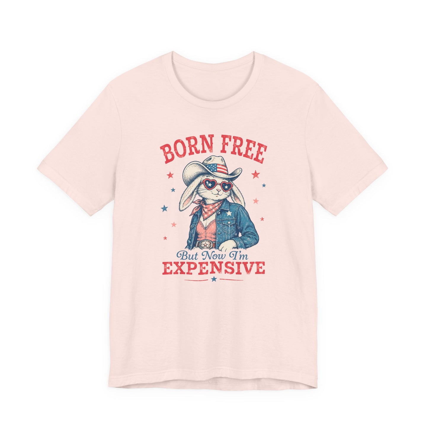 Adult Bella + Canvas light pink t-shirt featuring a patriotic bunny cowgirl graphic with heart sunglasses, American flag cowboy hat, denim jacket, and the phrase “Born Free But Now I’m Expensive” in distressed red and blue lettering.