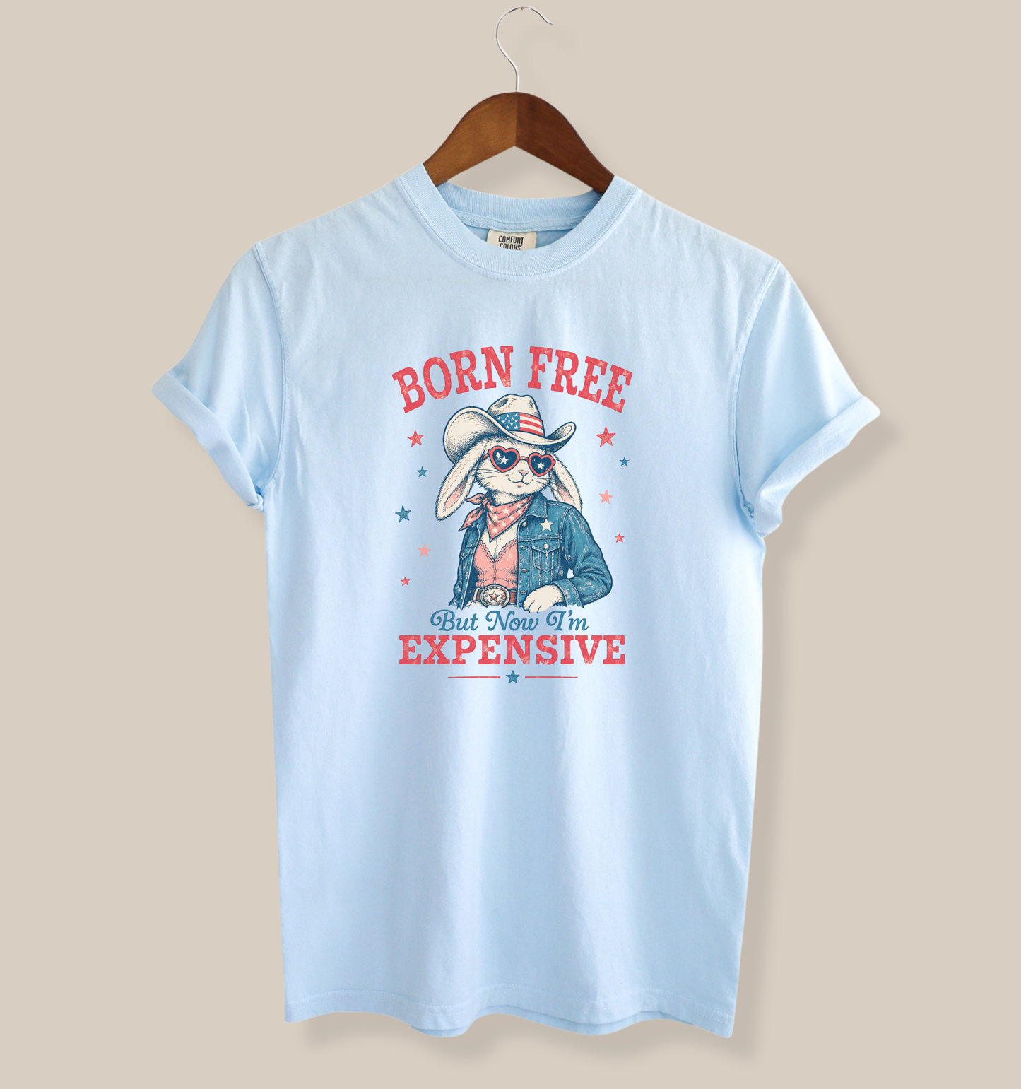 Adult Comfort Colors chambray blue t-shirt featuring a patriotic bunny cowgirl graphic with heart sunglasses, denim jacket, cowboy hat, and the phrase “Born Free But Now I’m Expensive” in vintage red and blue lettering.
