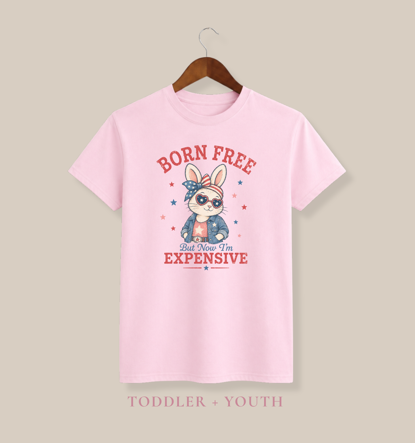 Bella + Canvas youth soft pink t-shirt featuring a patriotic bunny graphic with heart sunglasses, stars and stripes bow, denim jacket, and the phrase “Born Free But Now I’m Expensive” in vintage red and blue lettering.
