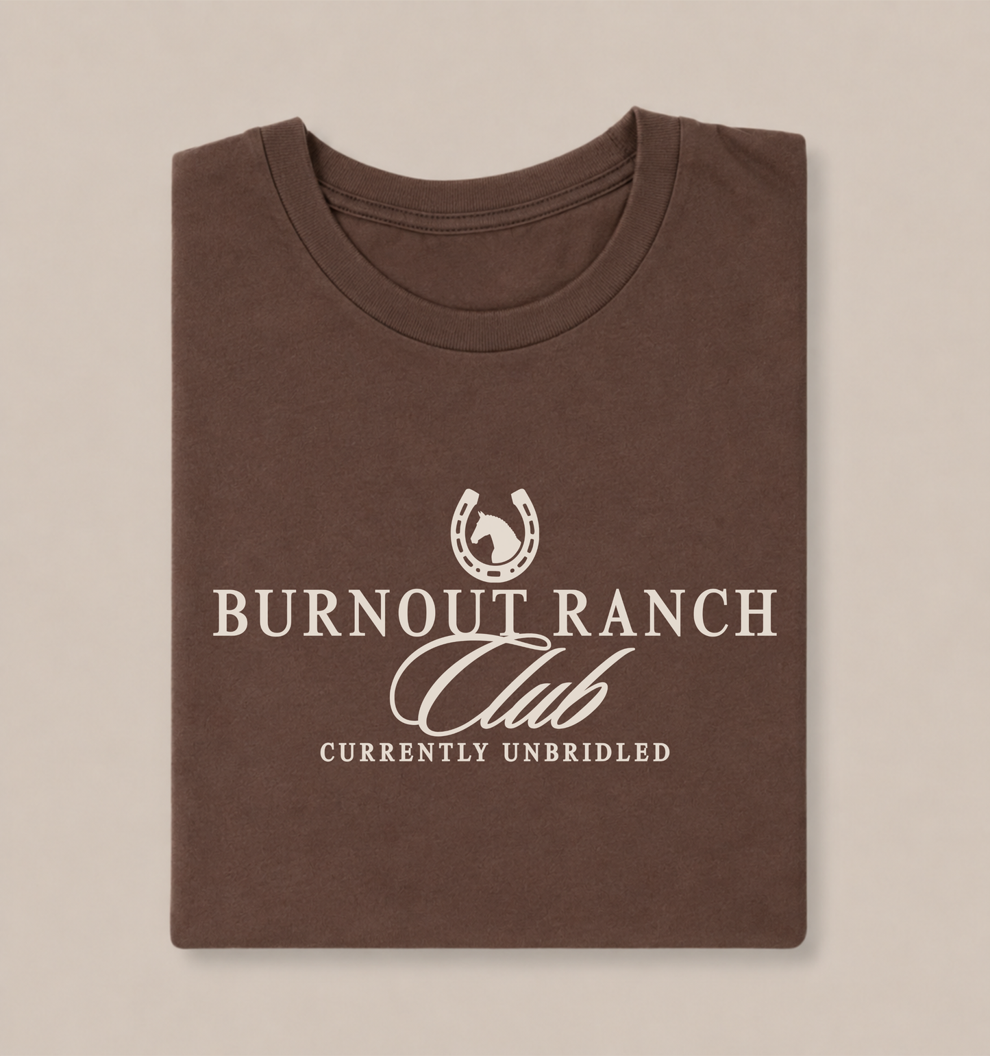 Folded brown Bella + Canvas 3001 t-shirt featuring a western-inspired “Burnout Ranch Club” graphic with a horseshoe icon and the phrase “Currently Unbridled” in cream lettering.