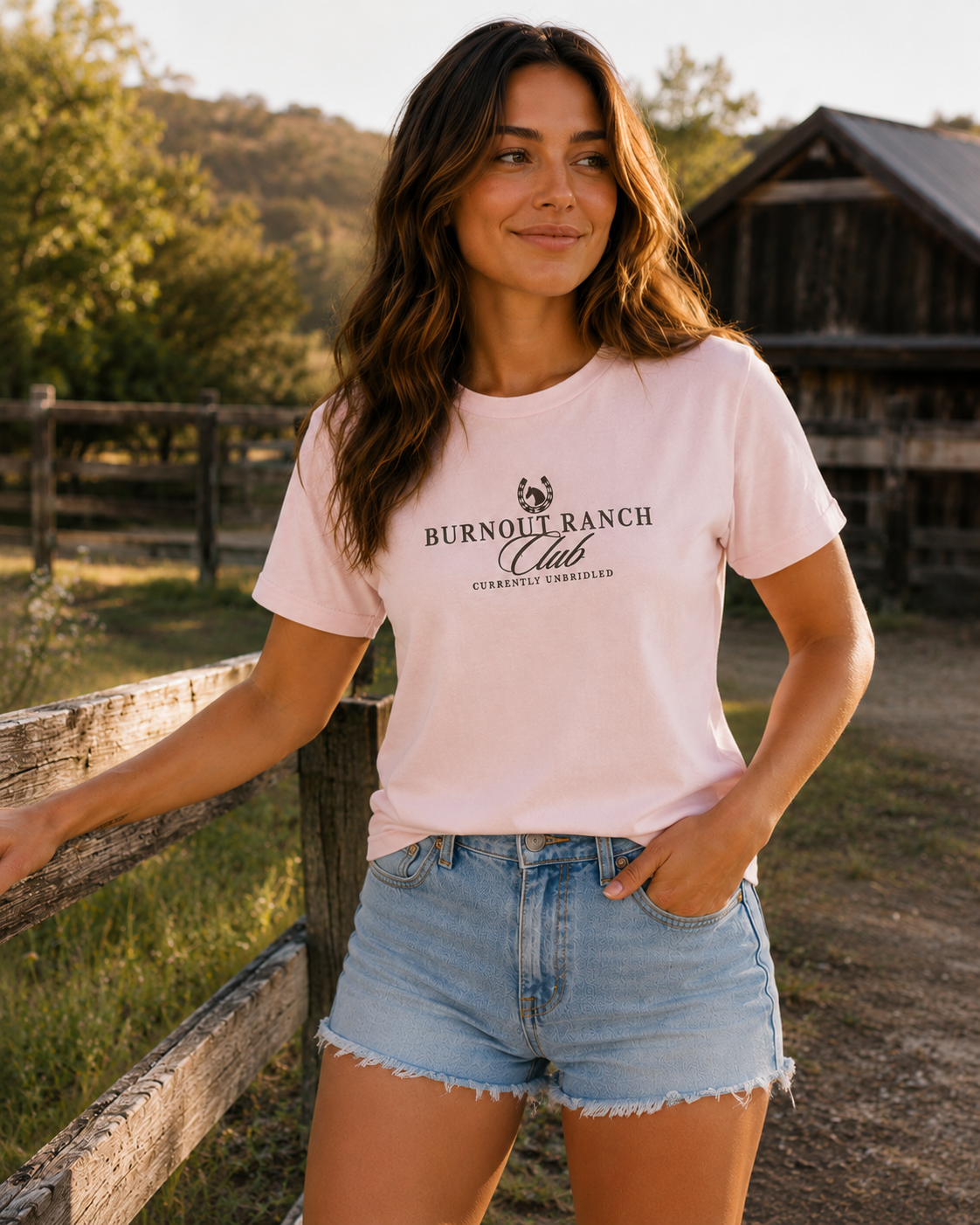 Woman wearing a soft pink Bella + Canvas 3001 t-shirt with a western-inspired “Burnout Ranch Club” graphic and “Currently Unbridled” text while standing at a rustic ranch during golden hour.