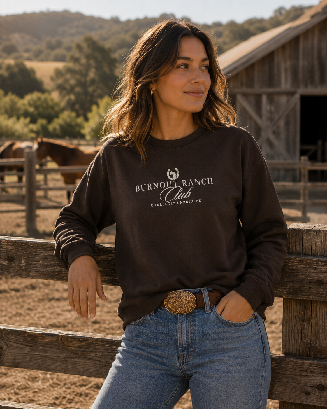 Woman wearing a brown Gildan 18000 sweatshirt with cream “Burnout Ranch Club” graphic and “Currently Unbridled” text while standing at a rustic horse ranch near a wooden fence and barn.